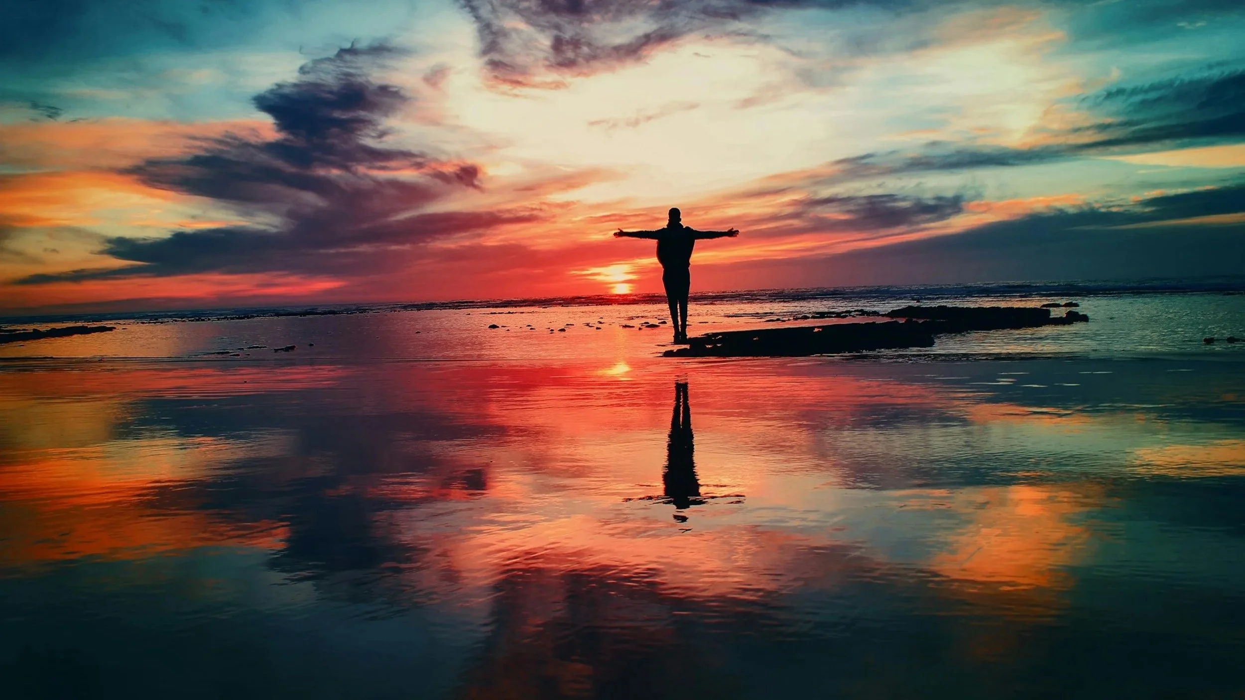 A person standing on a rock formation in the water with arms outstretched during sunset, reflecting in the calm water.