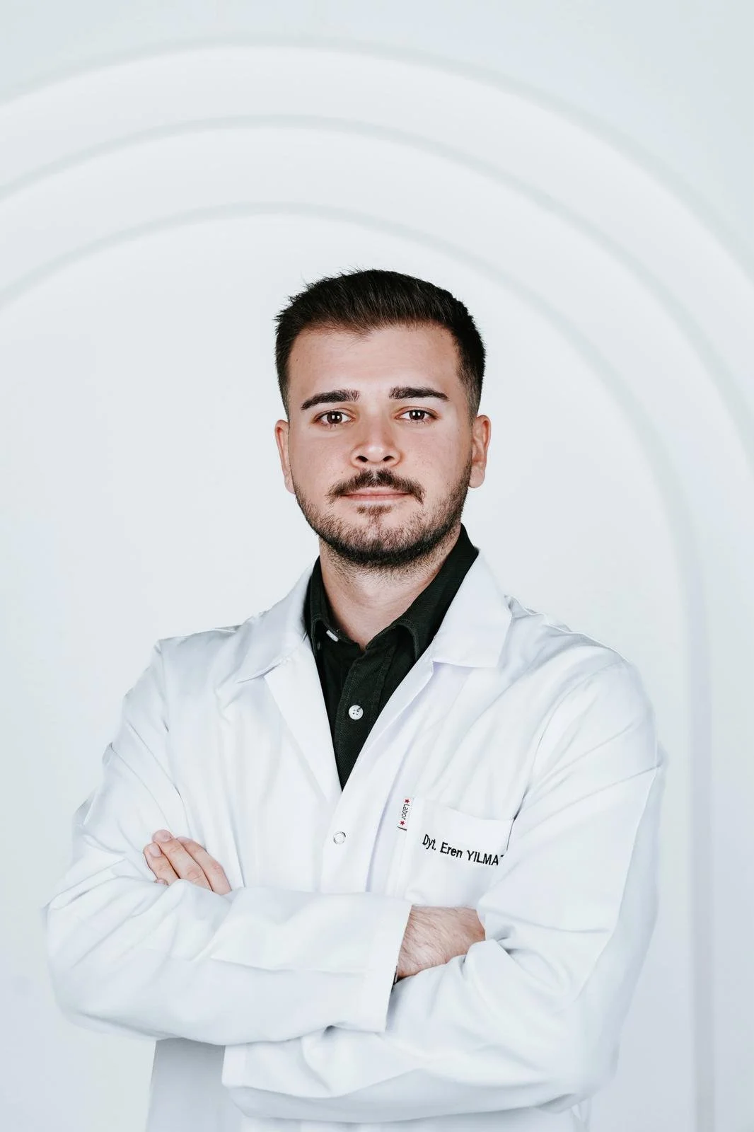 Beyaz önlük giymiş genç erkek diyetisyen, kolları çapraz ve arka planda açık renkli, nötr bir fon ile duruyor.