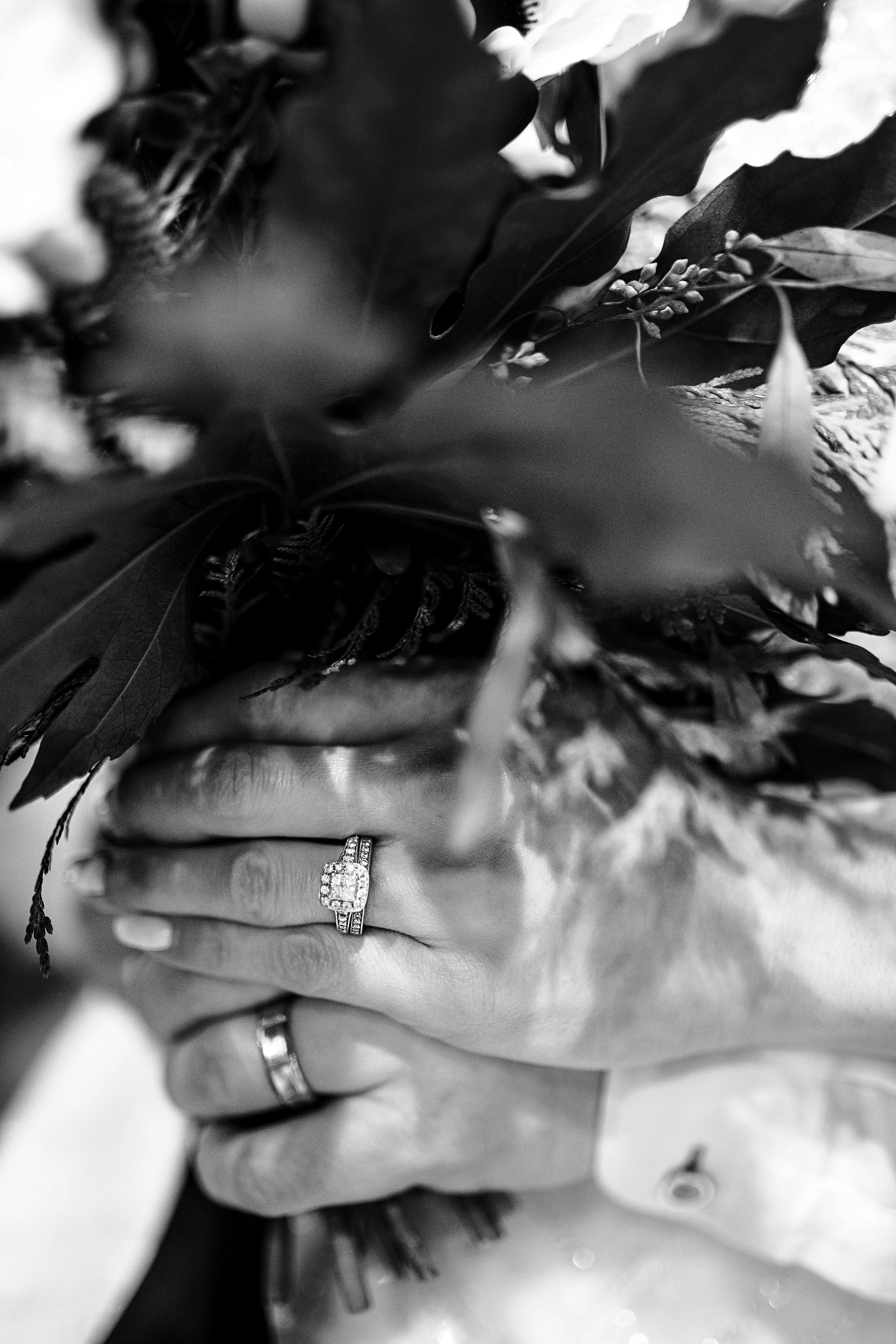 Close-up of a woman's hand with rings, holding a bouquet of leaves and flowers, in black and white.
