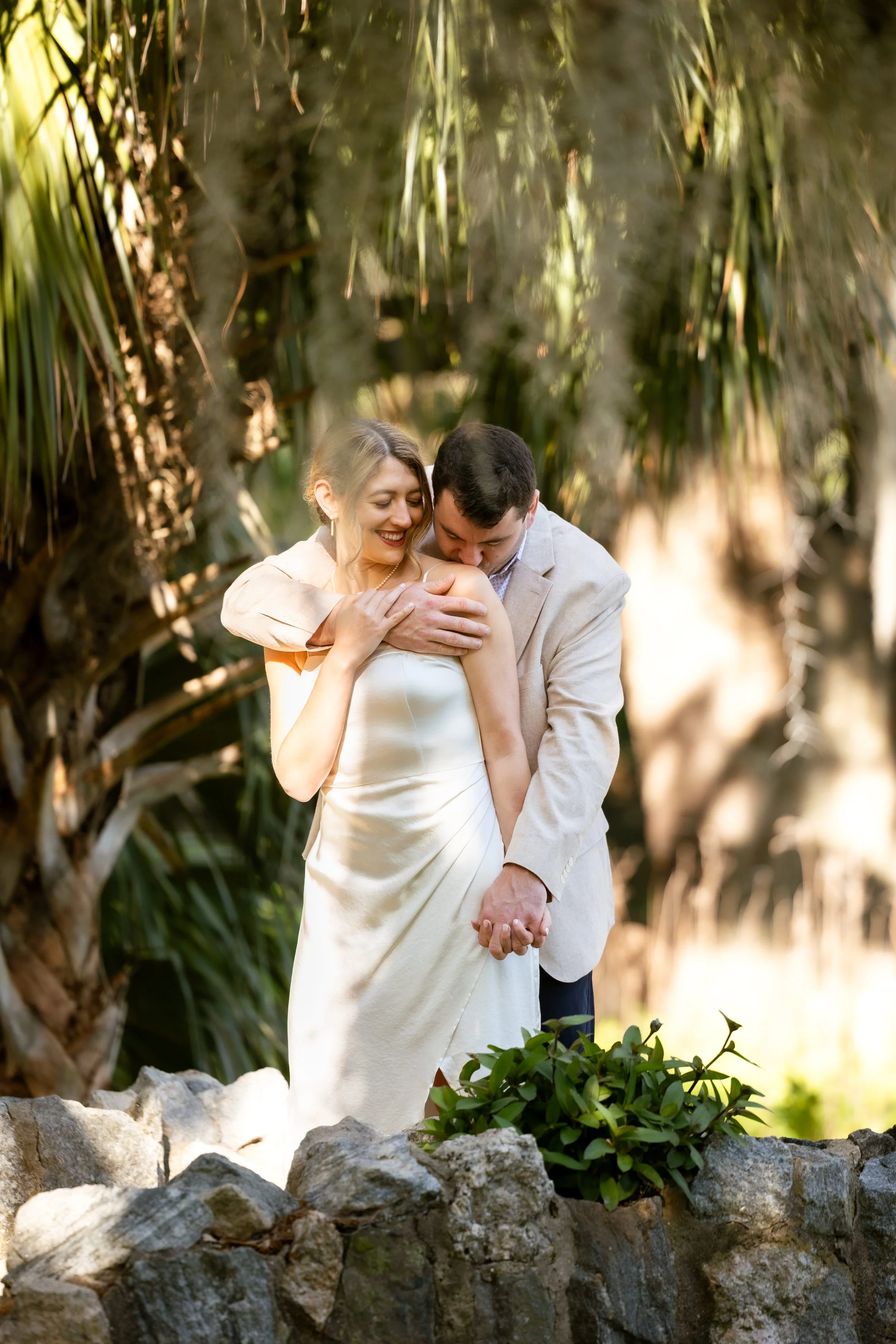 A couple dressed in wedding attire sharing an intimate moment outdoors, surrounded by greenery and rocks.