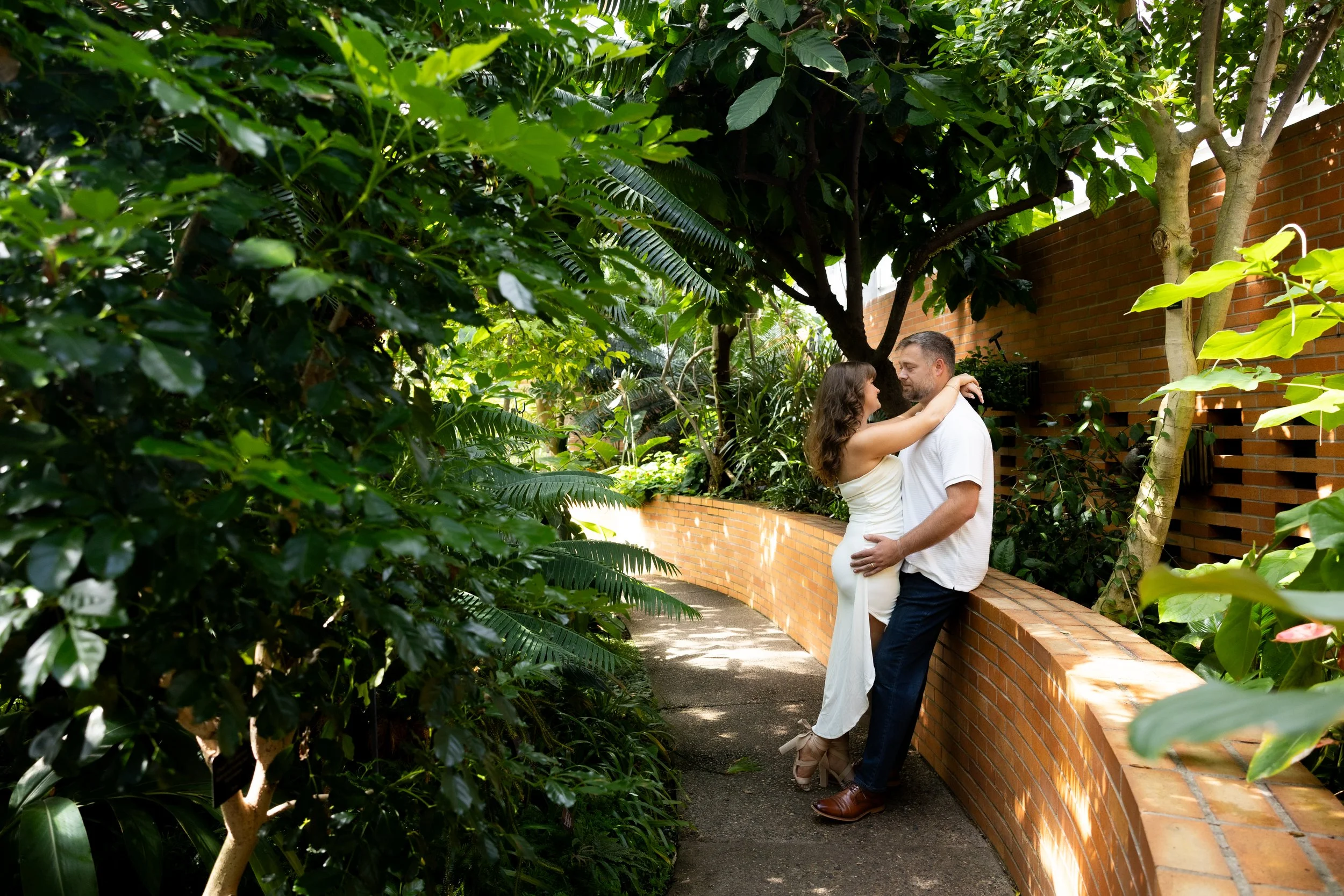 A couple embracing on a curved brick wall in a lush, green garden.