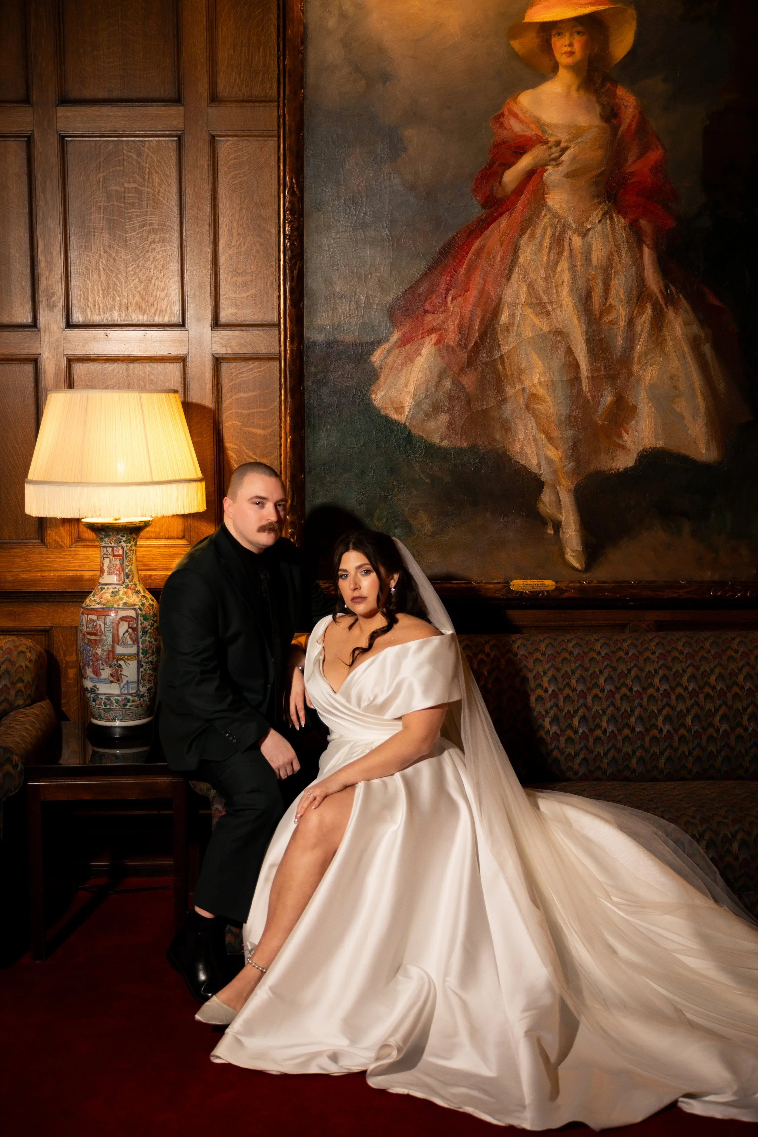 A bride and groom sitting on a vintage sofa in a dimly lit room with wooden paneling and a large portrait painting of a woman in historical attire hanging on the wall behind them.