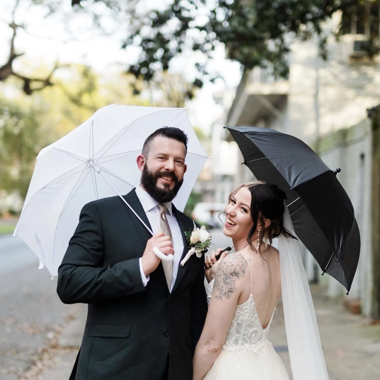 A bride and groom in wedding attire standing outdoors with umbrellas, smiling at the camera.