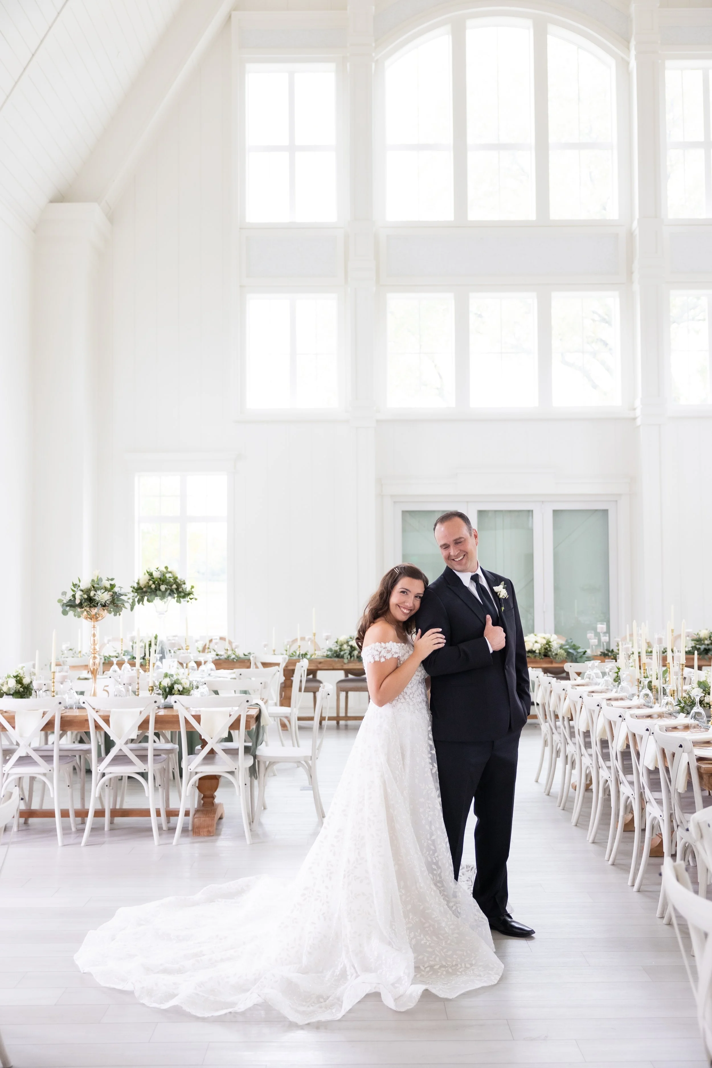 Bride in a white wedding gown and groom in a black tuxedo standing together inside a bright, elegant wedding venue decorated with white flowers and candles.