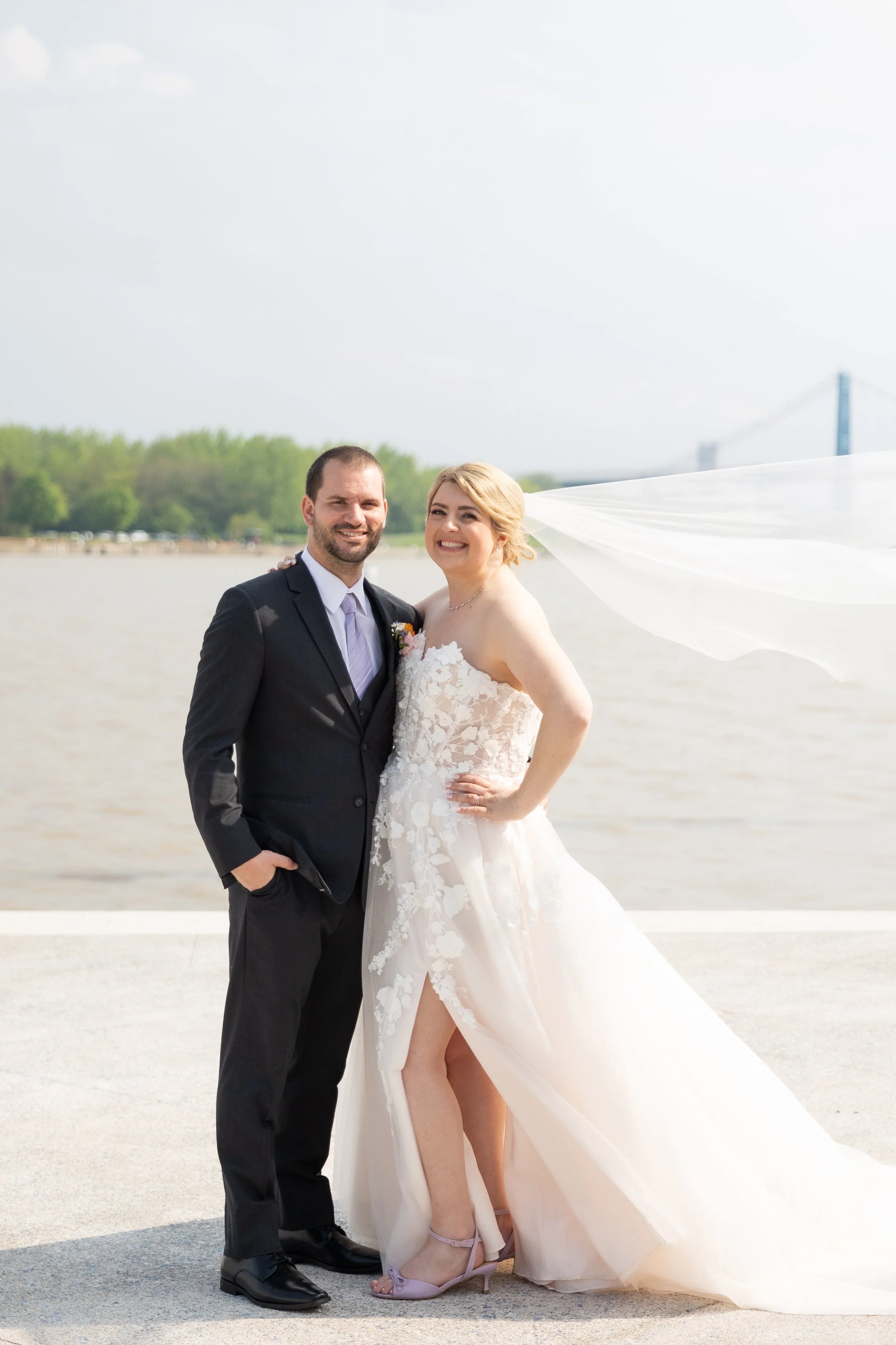 Bride and groom posing outdoors near a body of water, with a bridge in the background on a bright day.