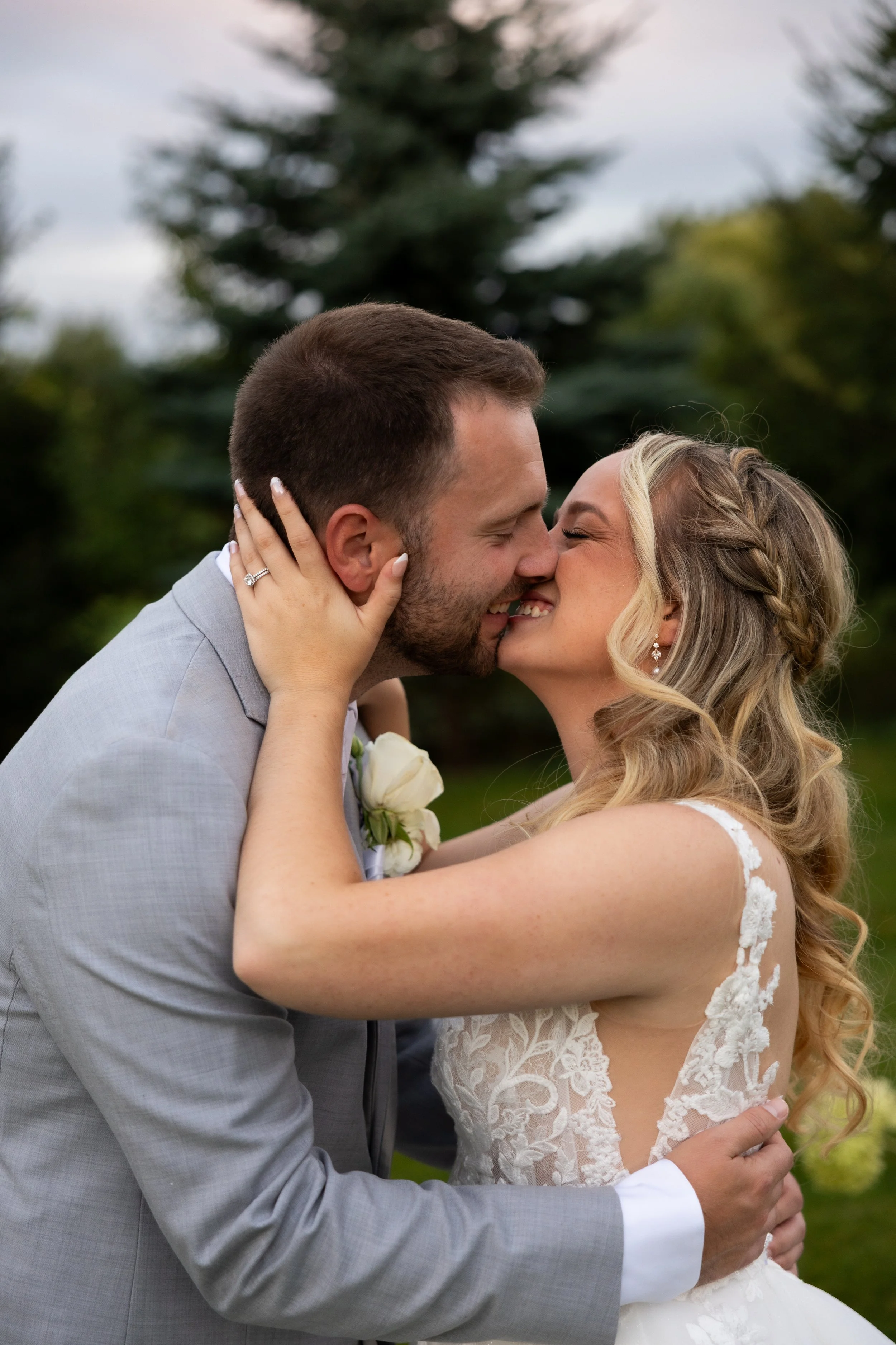 A newlywed couple sharing a kiss outdoors on their wedding day, with the bride in a lace wedding dress and the groom in a light gray suit.