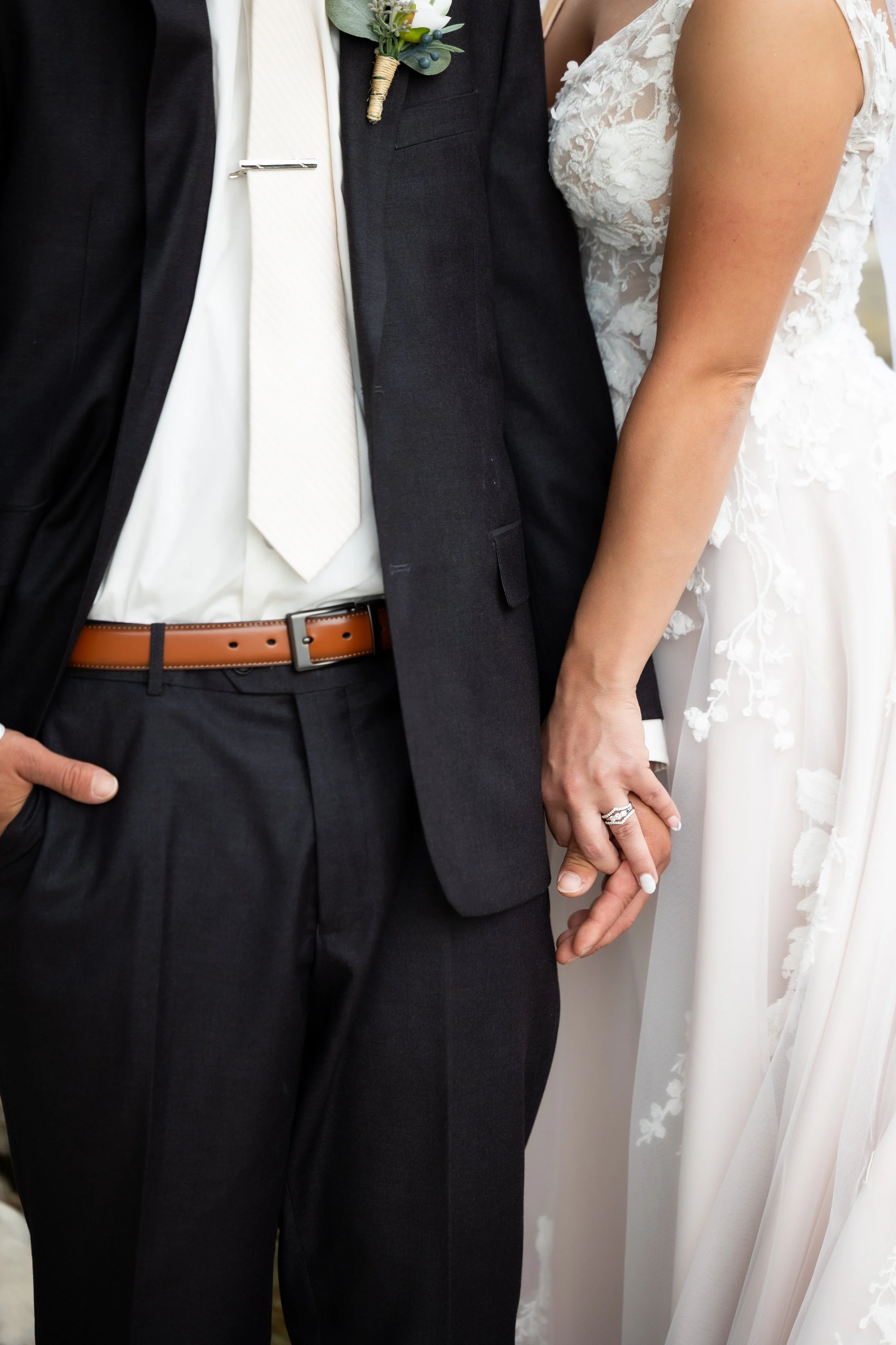 Close-up of a bride and groom holding hands during their wedding, with the bride wearing a white lace dress and a wedding ring, and the groom in a dark suit, white shirt, and brown belt.