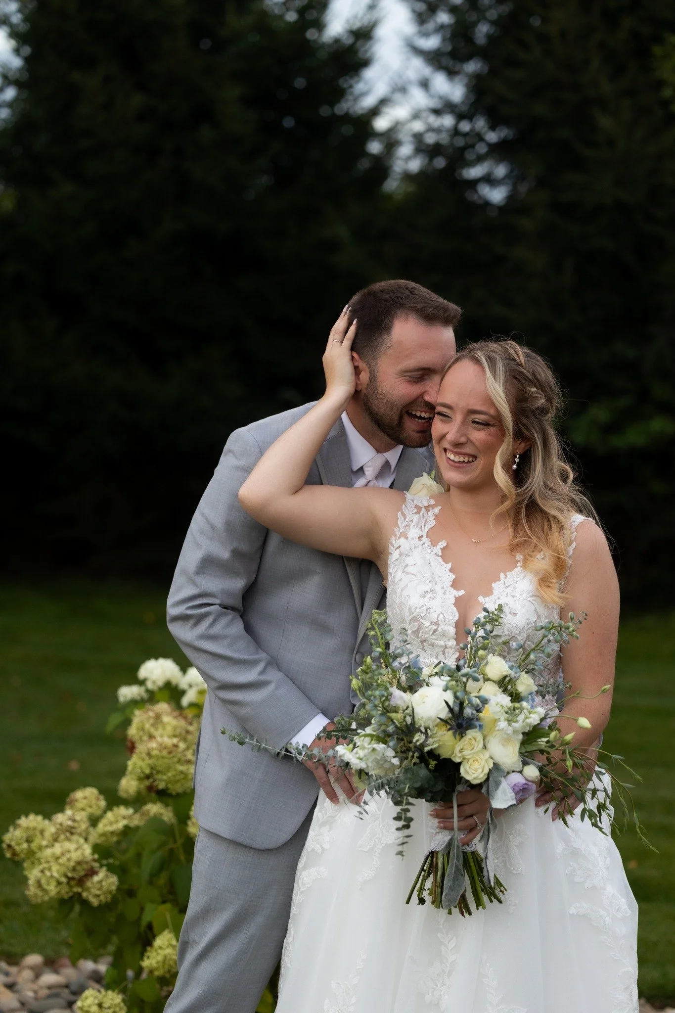 Happy bride and groom embracing outdoors on their wedding day, smiling with flowers in the bride's hand.
