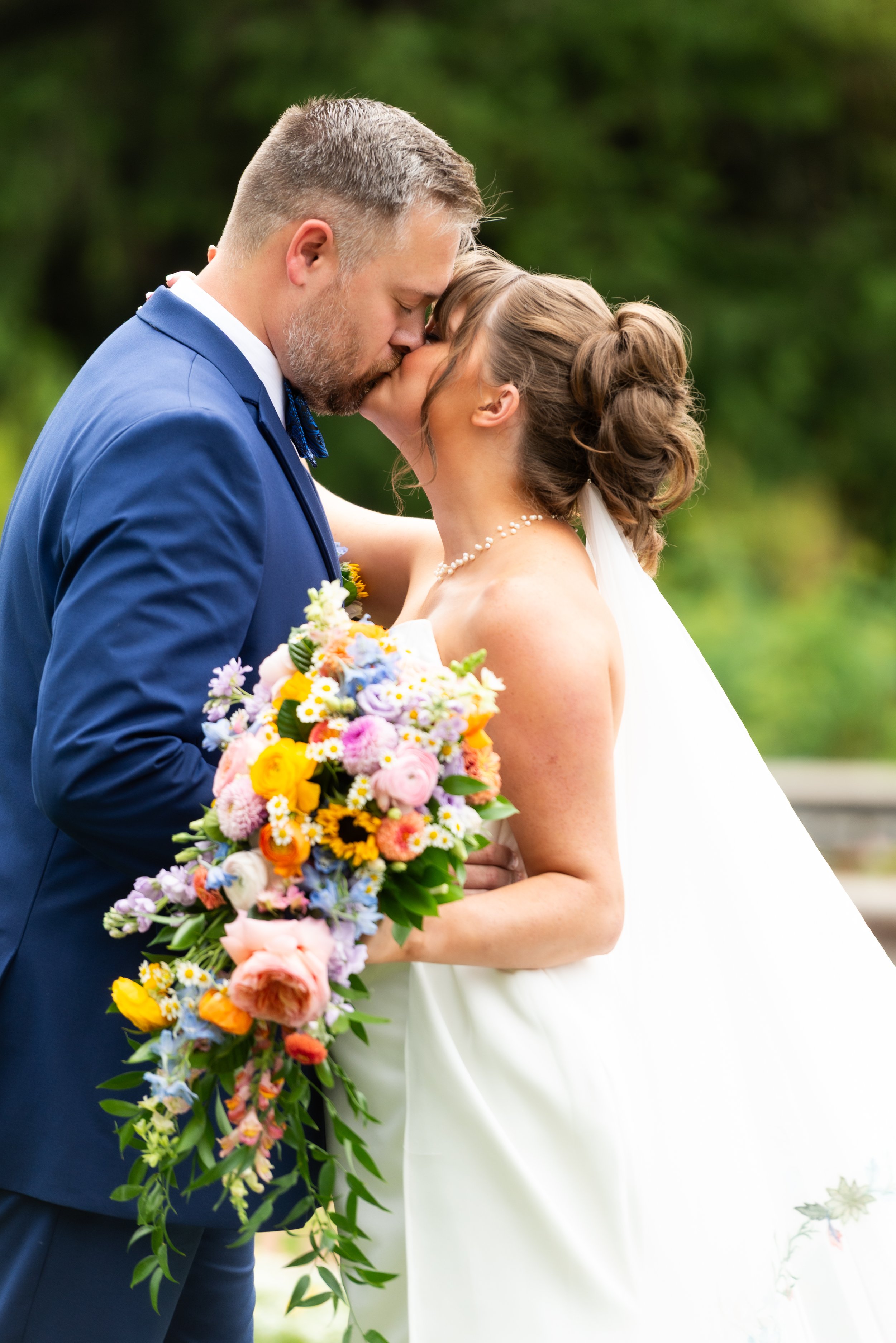 A bride and groom sharing a kiss outdoors, with the groom holding a colorful bouquet of flowers.