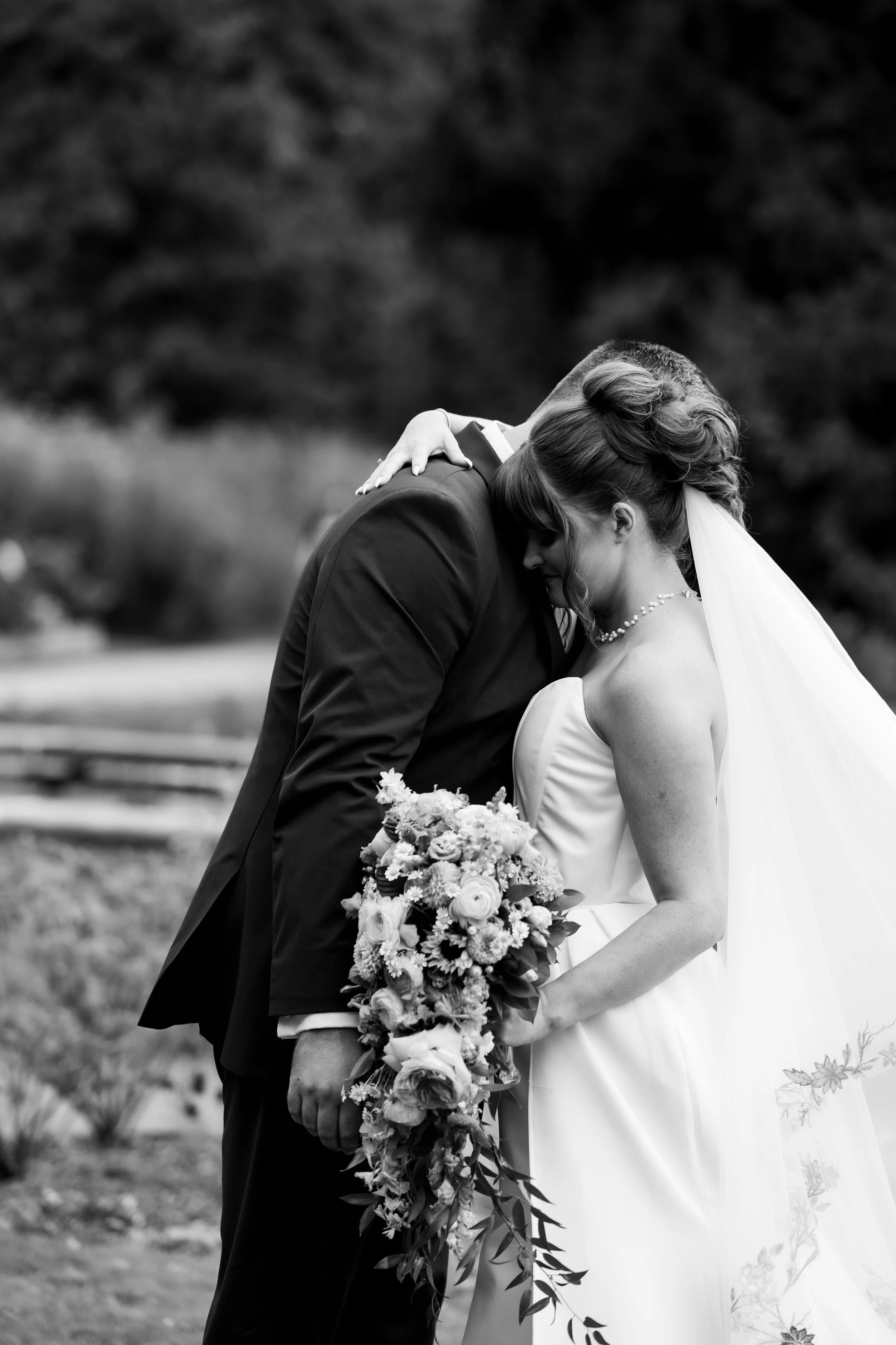 A black and white photo of a bride and groom embracing, with the groom in a suit and the bride in a wedding dress holding a bouquet of flowers.