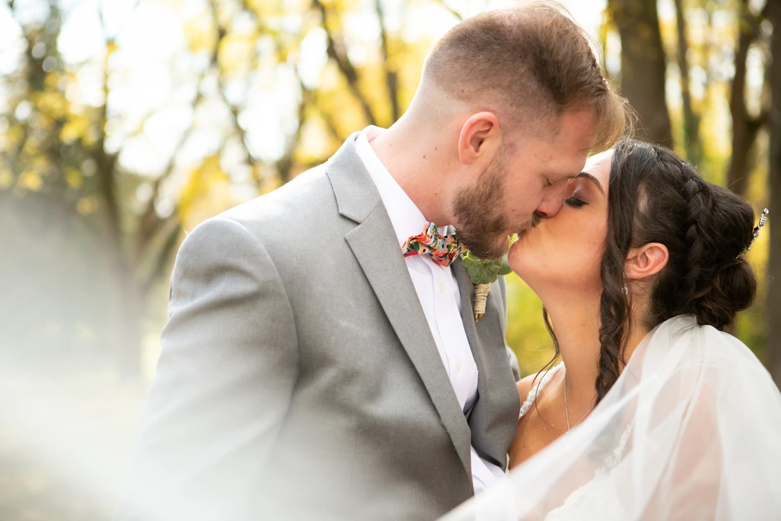 A couple in wedding attire standing close in a field of tall grass during sunset, with the bride wearing a white dress and wide-brimmed hat, and the groom in a plaid suit.