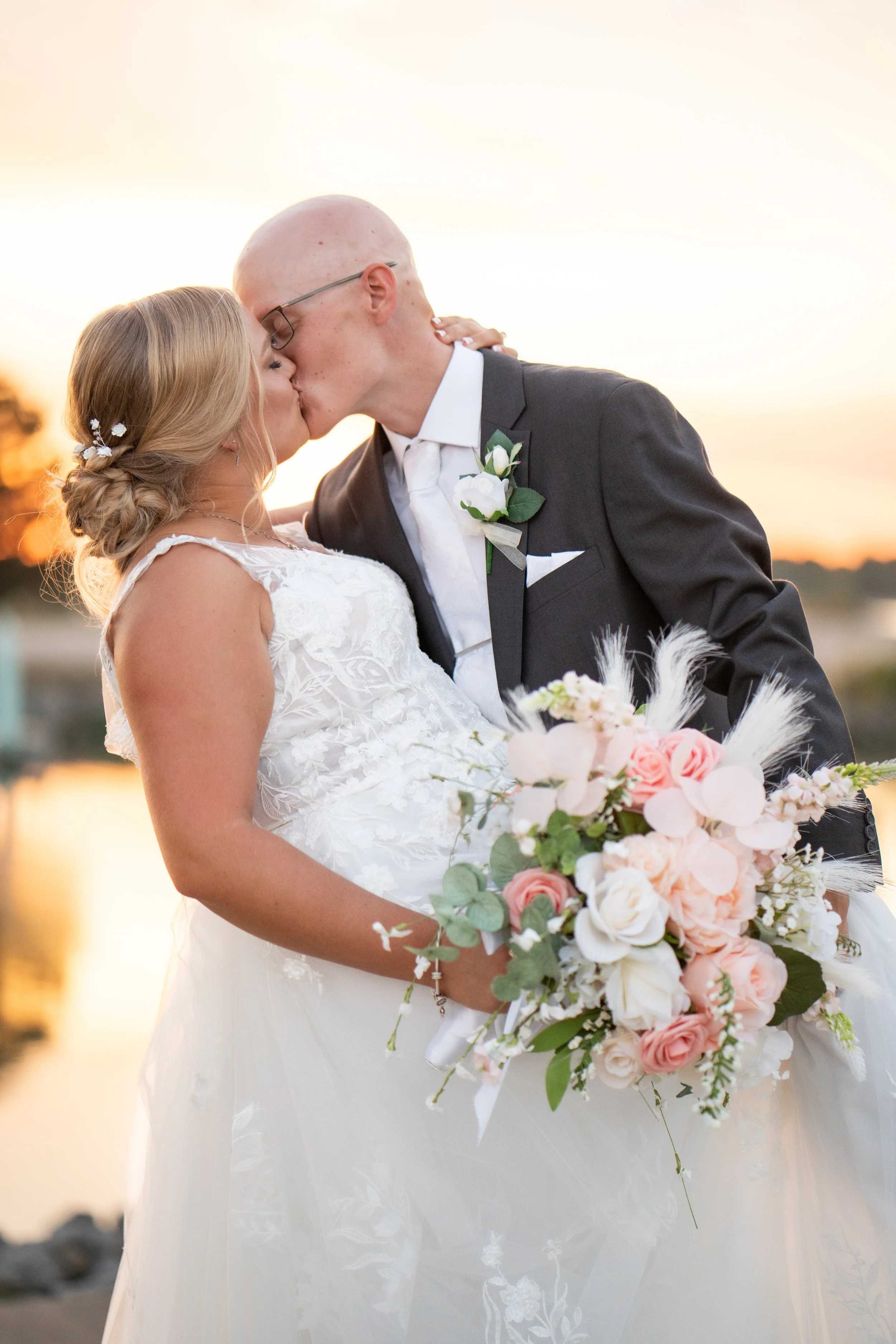 A newlywed couple sharing a kiss outdoors at sunset, with the bride holding a bouquet of pink and white roses and greenery, and the groom dressed in a black suit with a boutonniere.