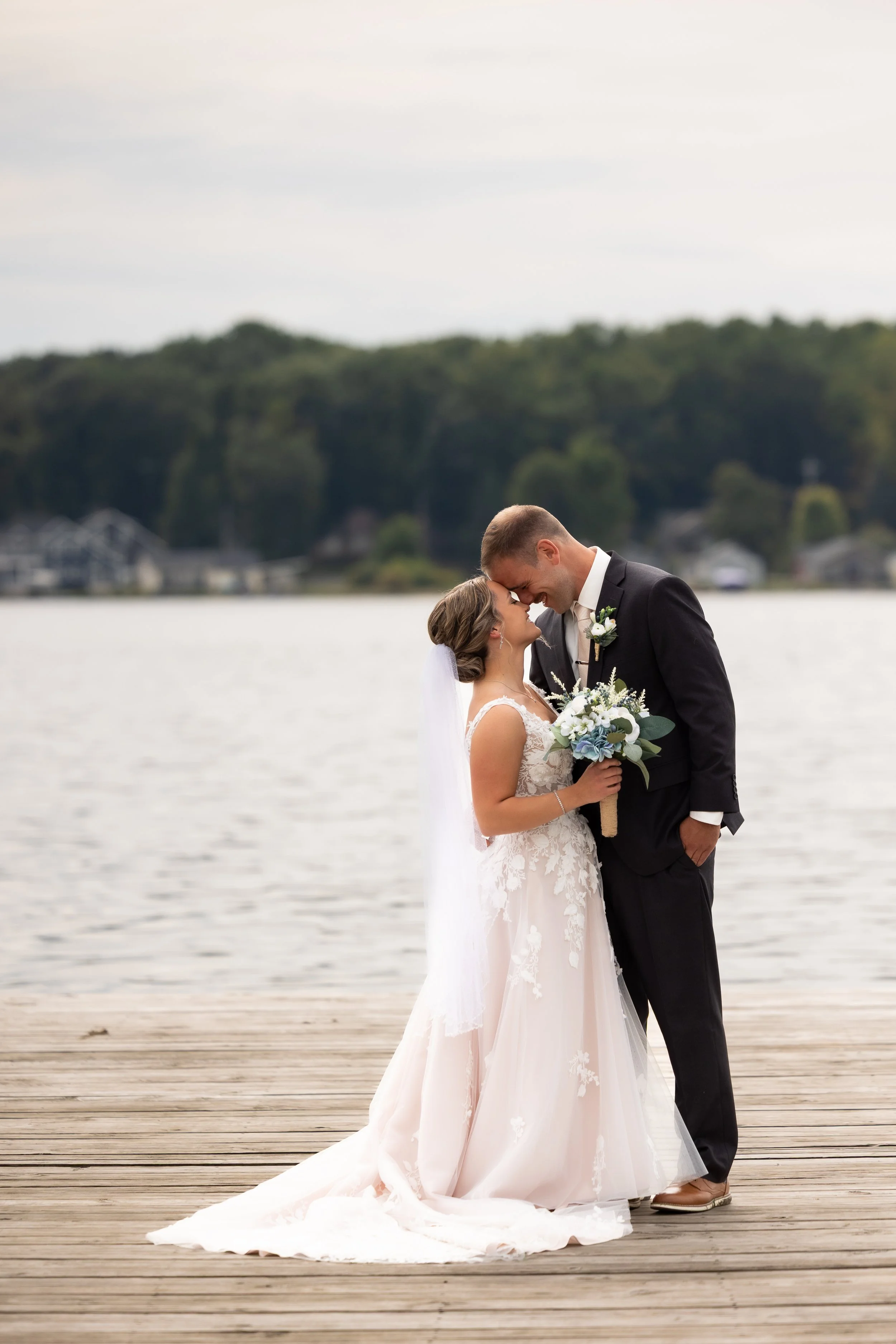 A bride and groom standing close together on a wooden dock by a lake, smiling with foreheads touching, during their wedding. The bride is wearing a white lace wedding dress and holding a bouquet of white and blue flowers, while the groom is dressed in a black suit with a white shirt and boutonniere. The background features water, trees, and a cloudy sky.