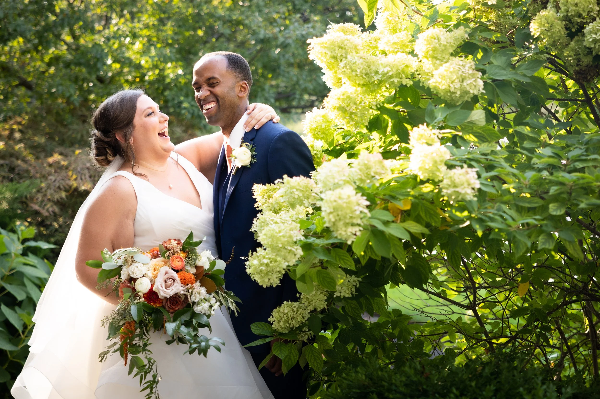 A newlywed couple, smiling and laughing, standing outside surrounded by green foliage and bright white flowers, with the bride holding a bouquet of flowers.