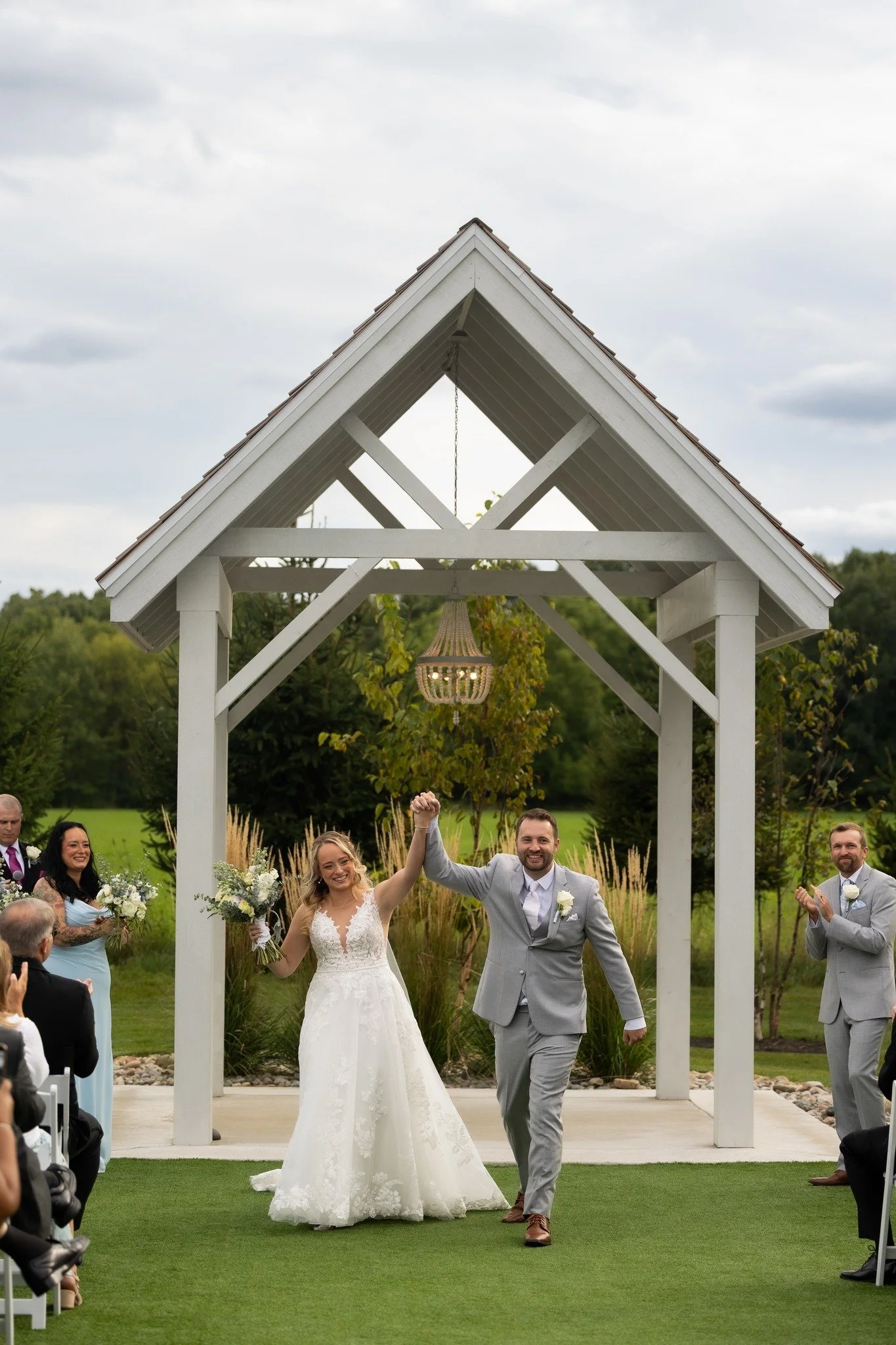 A newlywed couple walking down the aisle under a white wooden wedding arch, holding hands, with flowers in their other hands, while wedding guests celebrate around them outdoors.