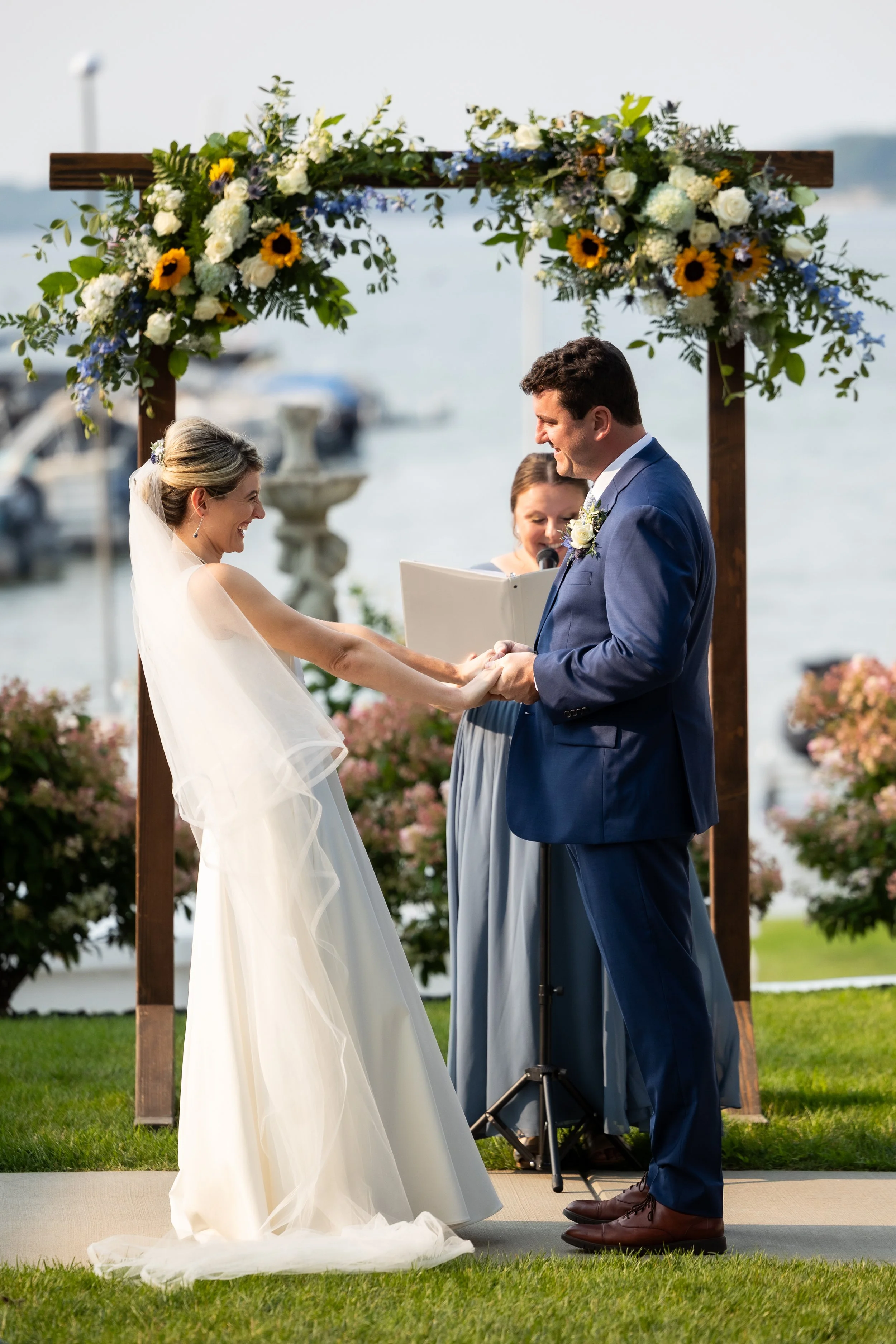 Wedding ceremony outdoors at a waterfront with a bride and groom holding hands under a floral arch, with an officiant reading from a book.
