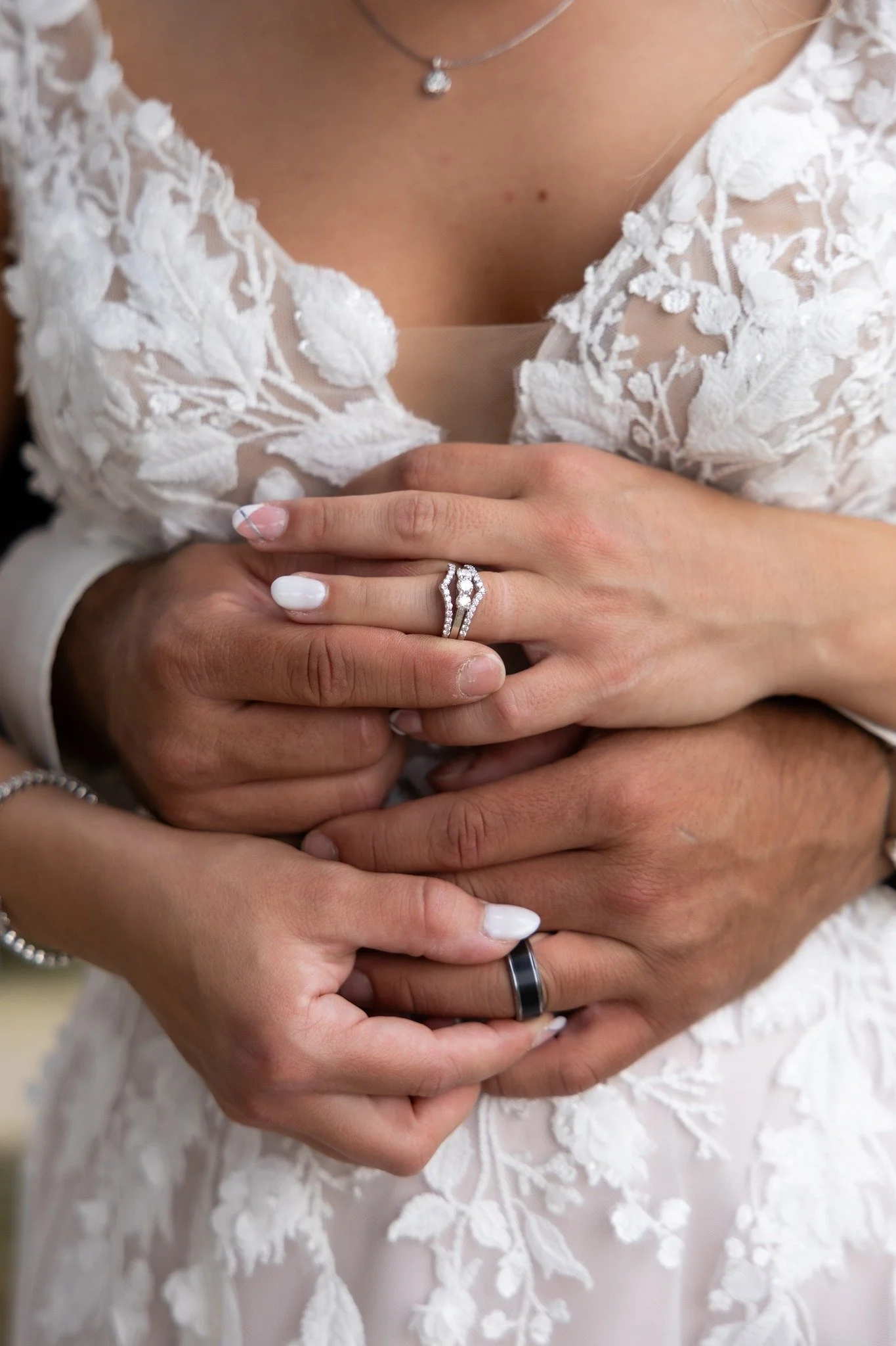 Close-up of two people holding hands, showcasing wedding rings; one person is wearing a white lacy wedding dress and a delicate necklace with a small pendant.
