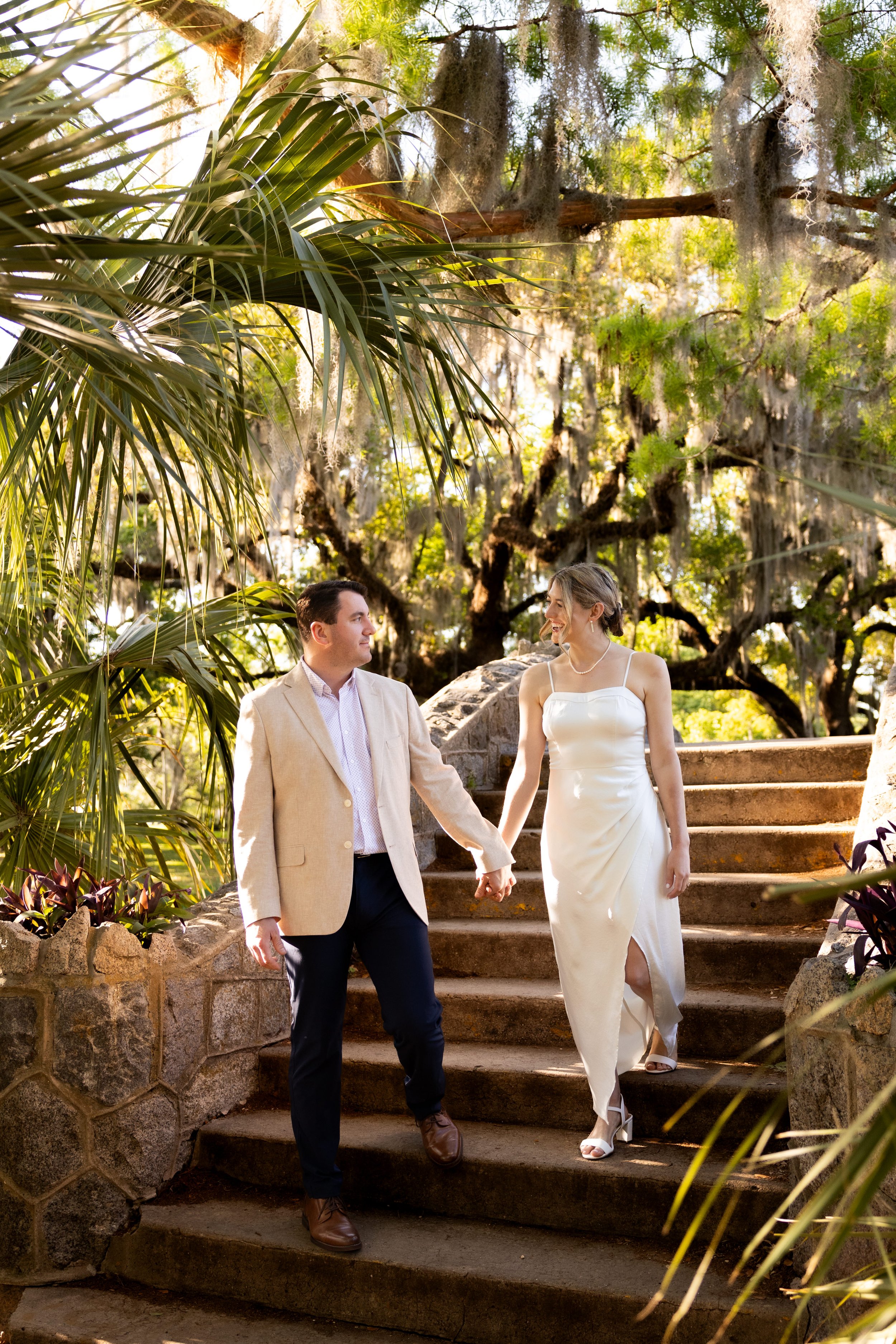 A couple holding hands walking down outdoor stone stairs surrounded by lush greenery and trees.