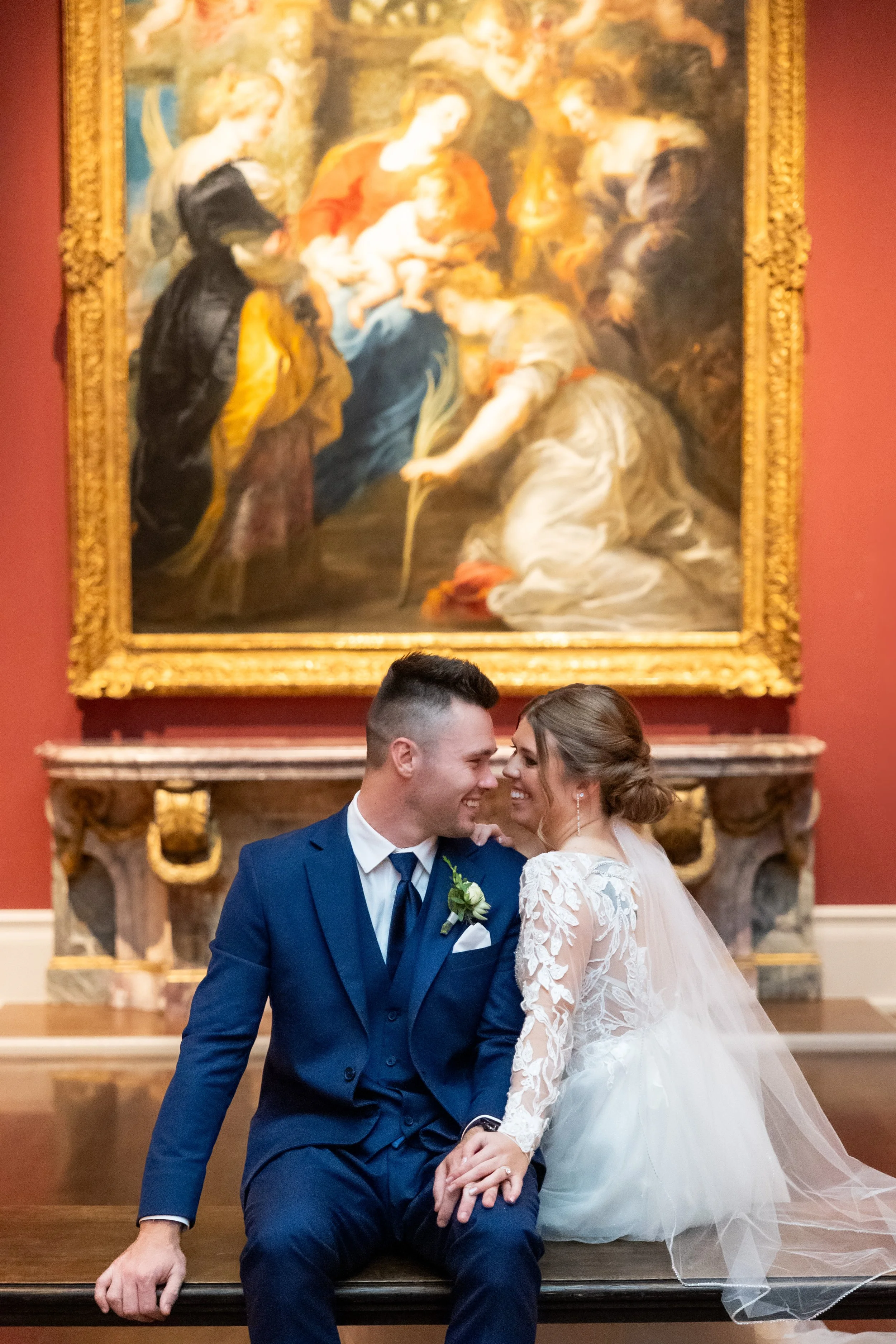 A bride and groom smiling and sitting close together on a bench inside an art museum, with a large classical painting in a gold frame behind them.