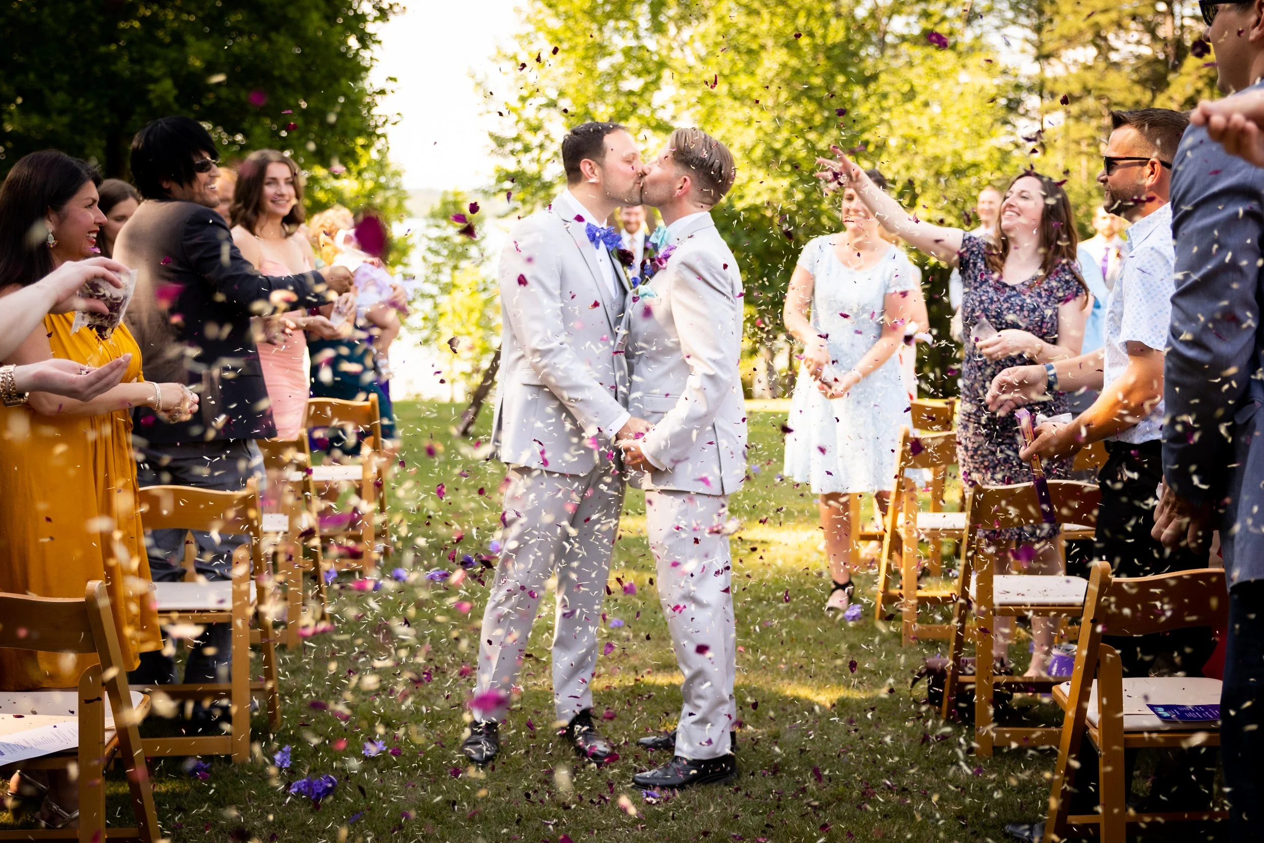 Two men in white suits kissing among friends and family at an outdoor wedding, surrounded by confetti and trees.
