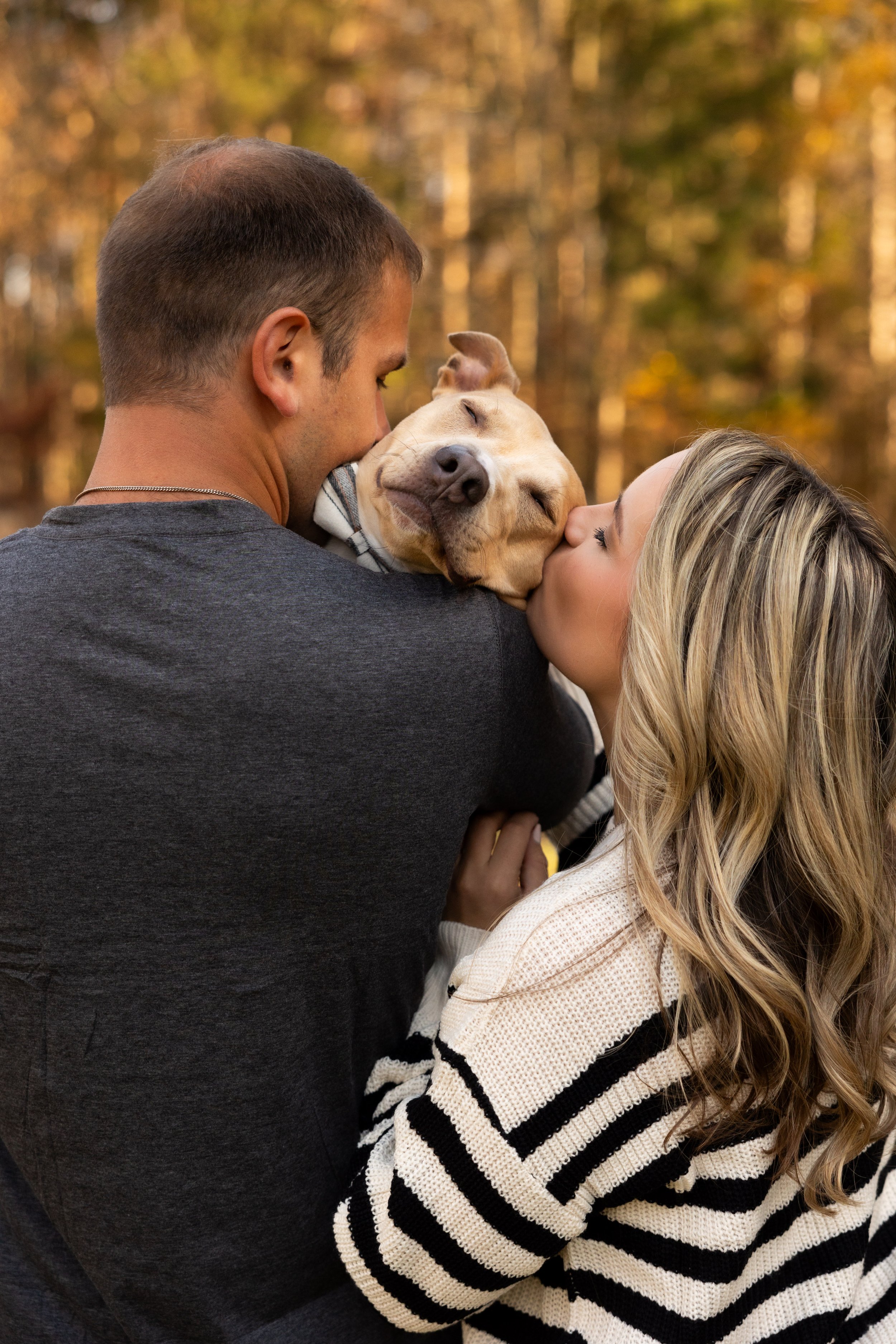 A man and woman kissing a smiling dog outdoors during fall, with trees in the background.