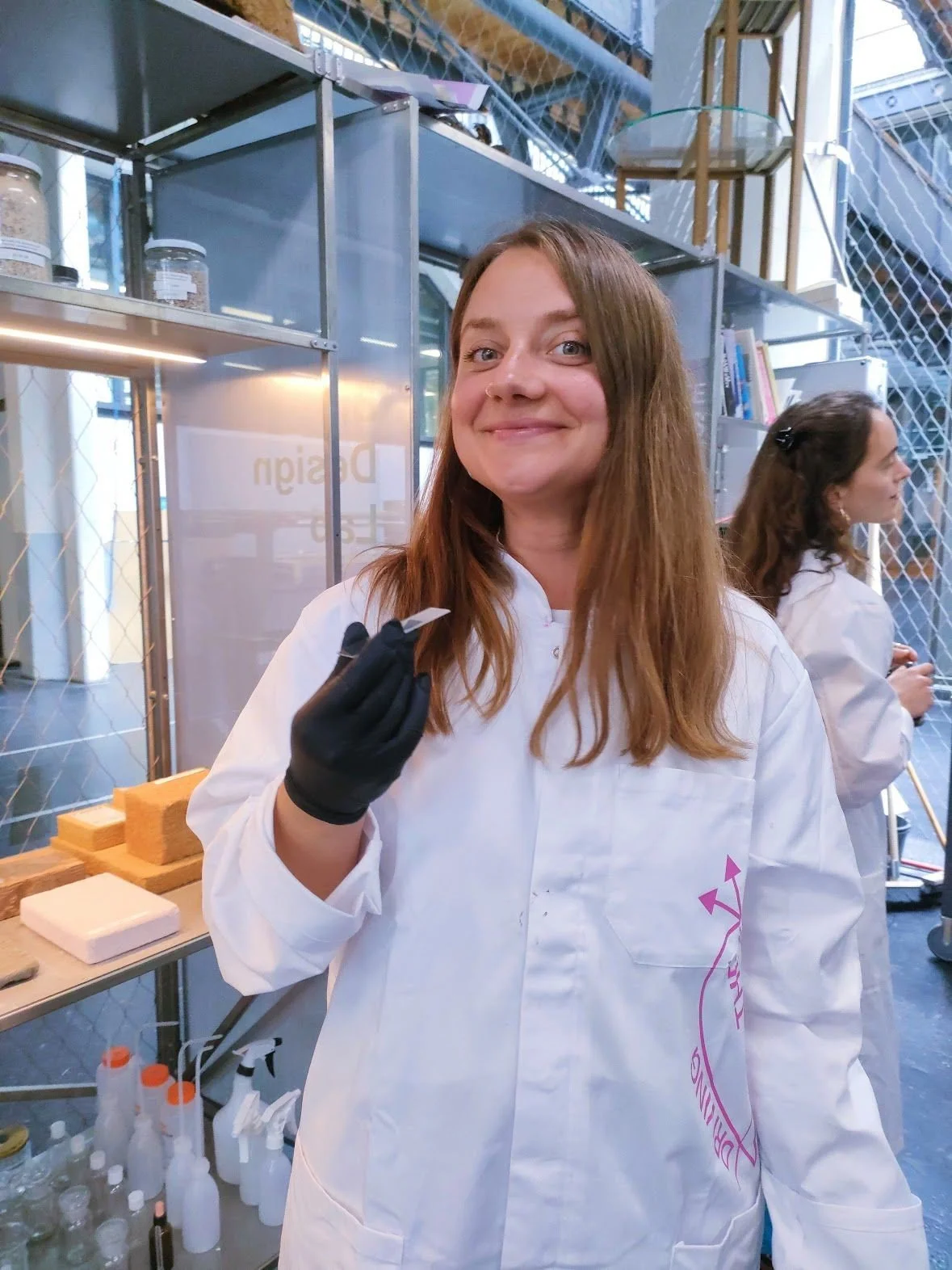 A woman with brown hair, wearing a white lab coat and black gloves, smiling at the camera inside a scientific laboratory or workspace.