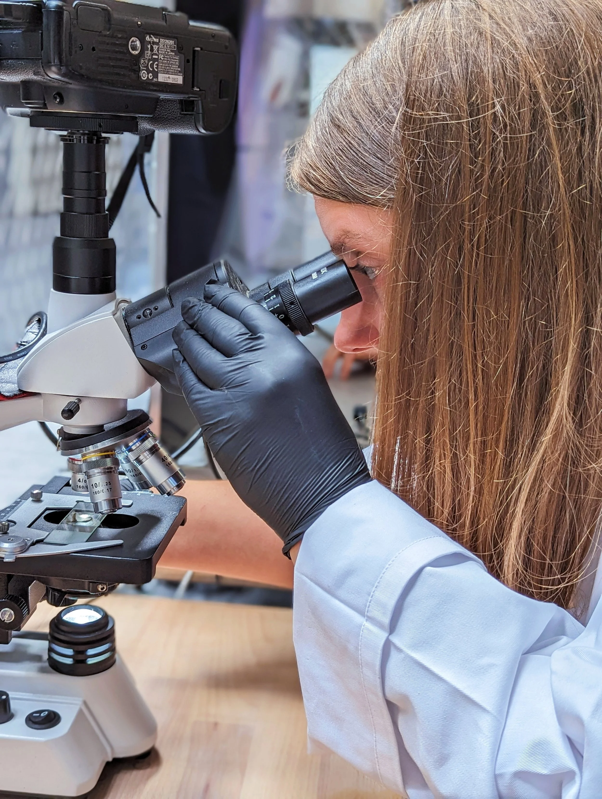 A scientist in a white lab coat and black gloves looking into a microscope in a laboratory setting.