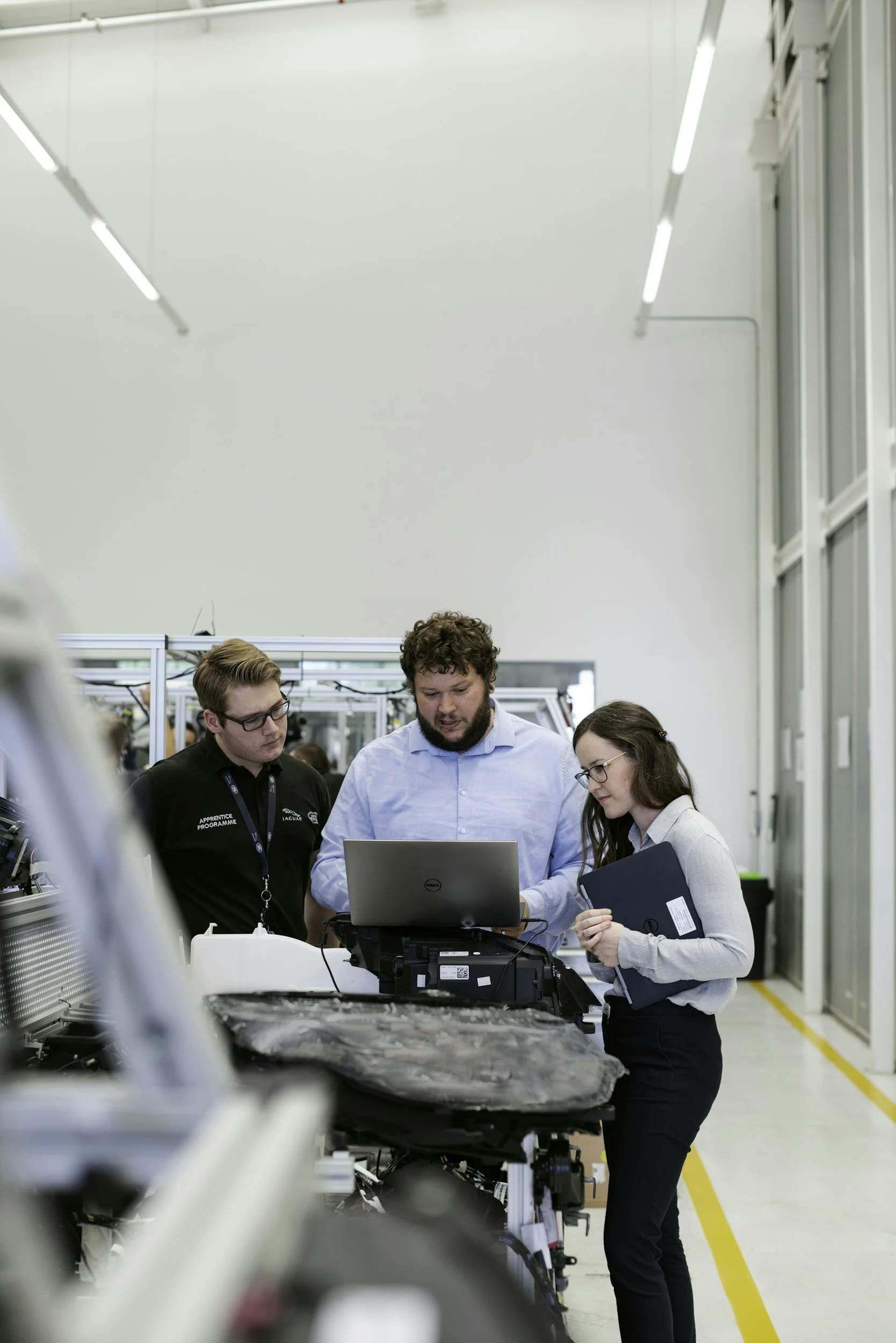 Three individuals examining machinery with a laptop and clipboard in a high-tech industrial or manufacturing facility.
