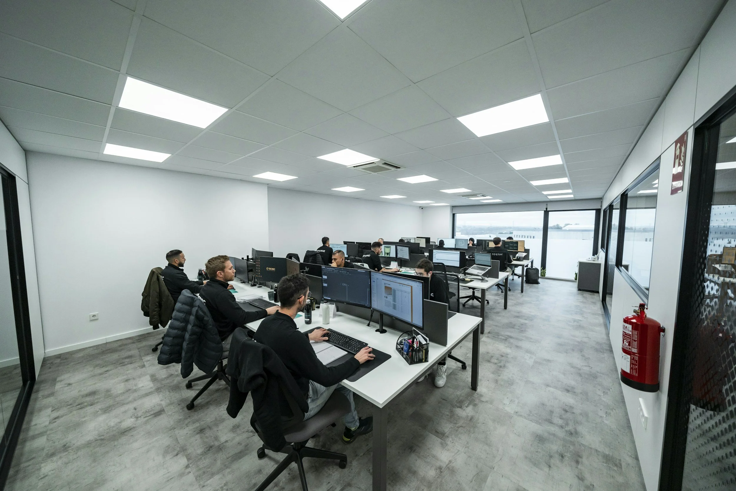 Office workers working at their desks in a modern office with large windows, white walls, and overhead lighting.