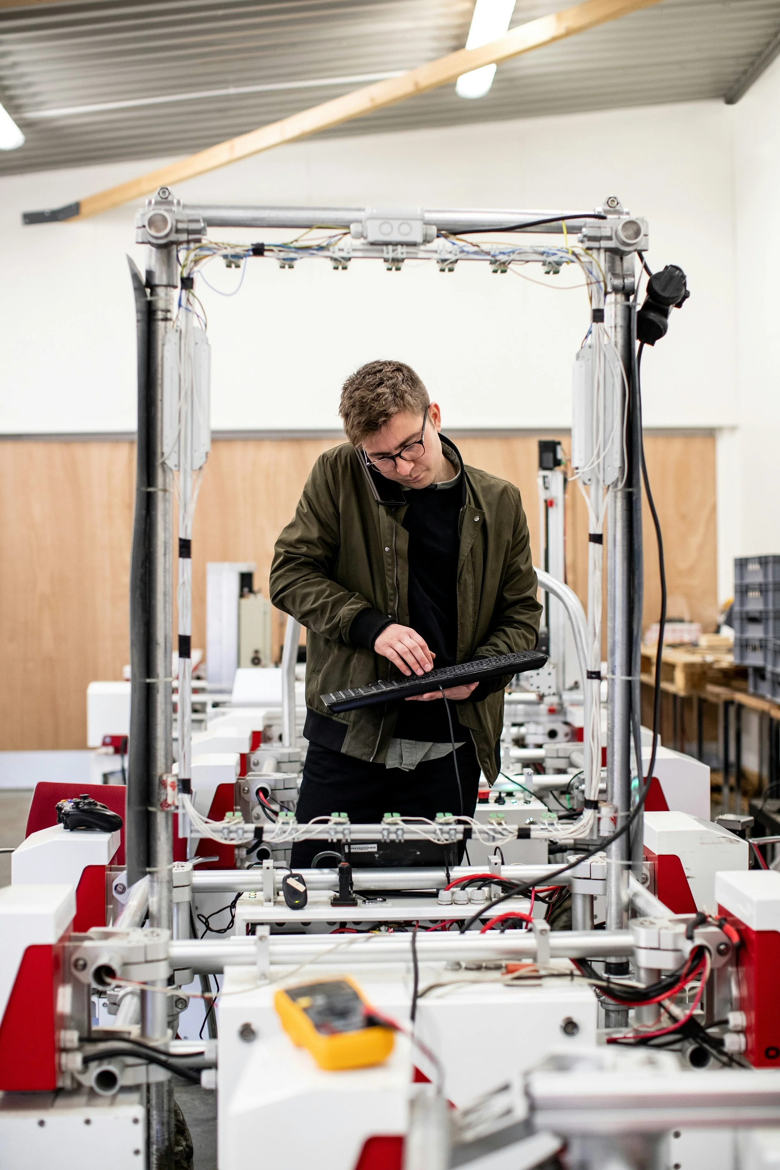 A young man with glasses wearing a green jacket working on a robot or machinery in a workshop, holding a keyboard and talking on a mobile phone.