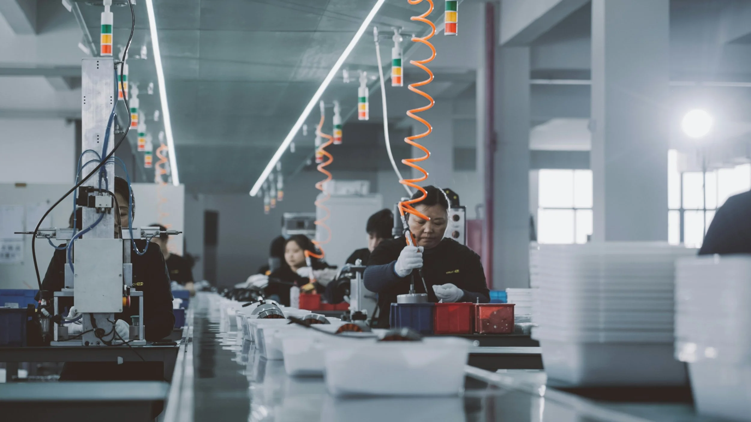 Workers assembling electronic devices on a factory production line, wearing gloves and working at their stations.