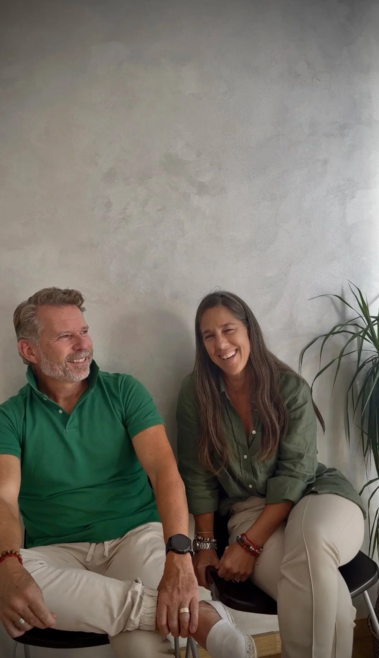 Una pareja riendo y sentada en una silla en una habitación con paredes de color claro, junto a una planta