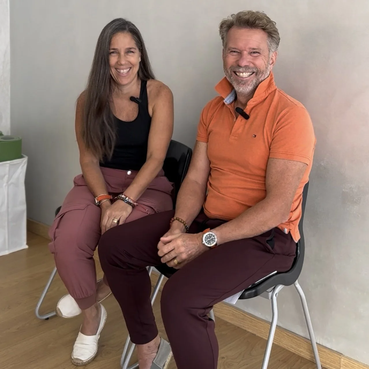 Dos personas, una mujer y un hombre, sentados en sillas frente a una pared blanca, sonriendo y usando micrófonos, probablemente en una entrevista o grabación.