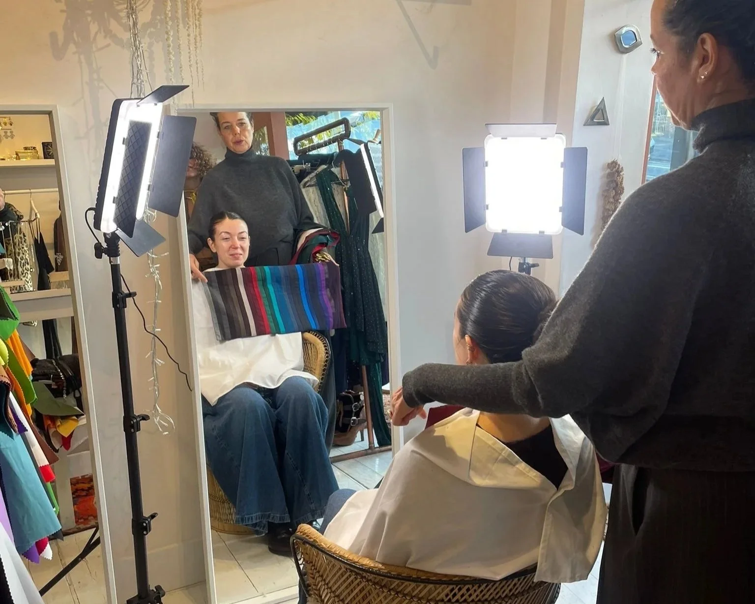 A woman sits in front of a mirror during a photoshoot with bright studio lights, with a stylist and a photographer assisting, all inside a boutique or studio with clothing displays.