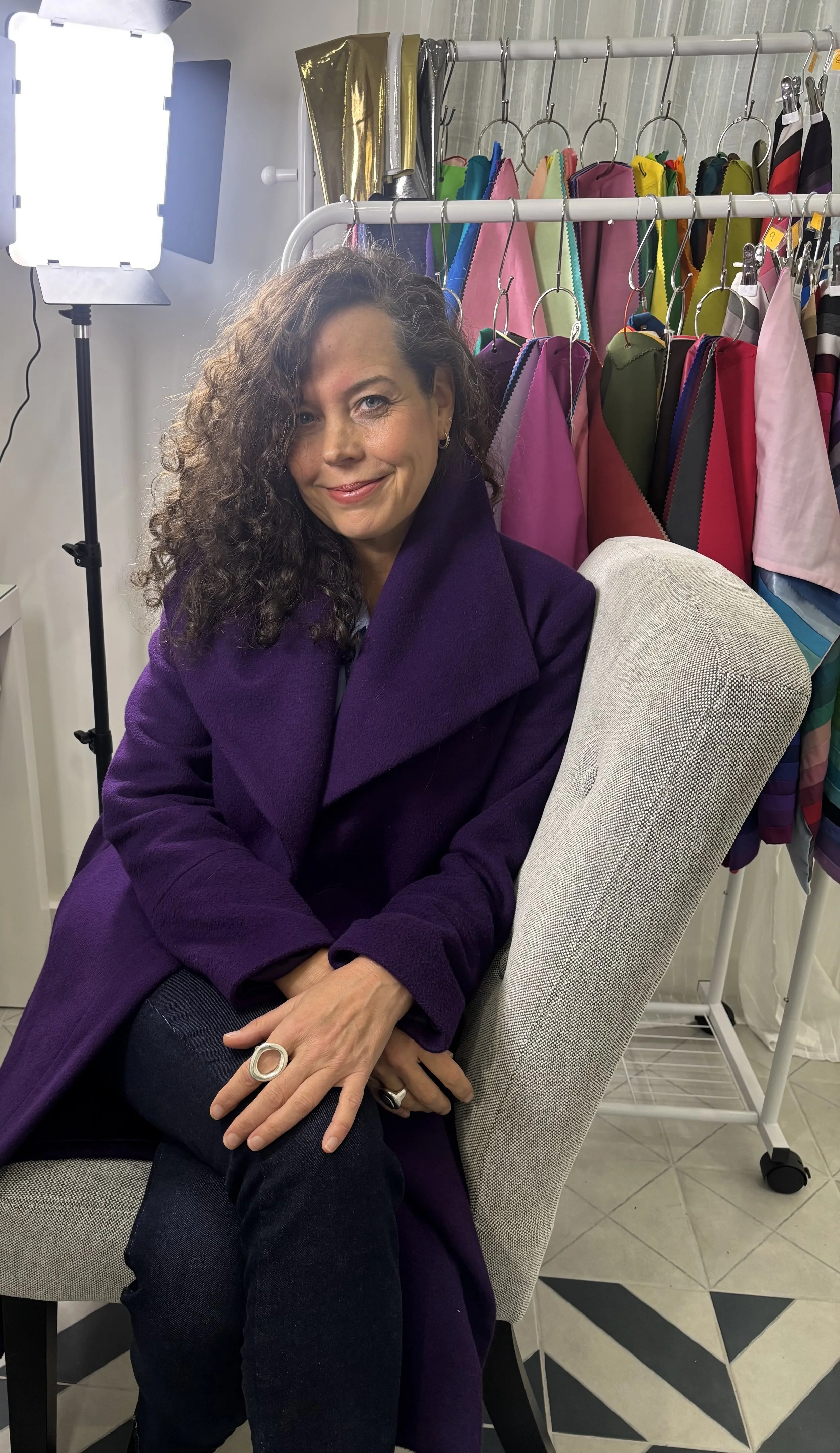 A woman with curly dark hair wearing a purple coat, sitting in a room with colorful fabric swatches hanging on a rack behind her. There is a lighting setup on her left.