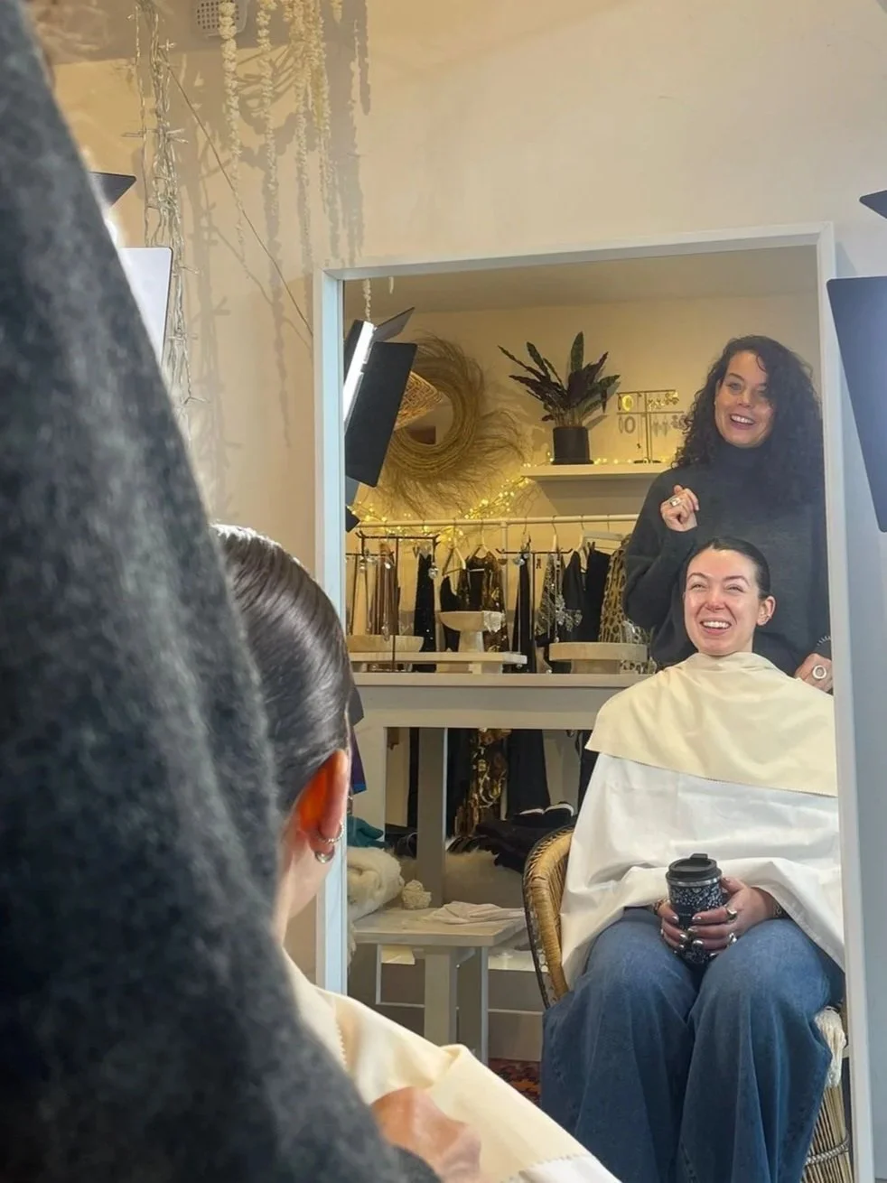 Woman in a hair salon getting styled, sitting in a chair with a cape, holding a travel mug, smiling at her reflection in a mirror, with a hairstylist standing behind her, inside a room with clothing racks and decorative plants.