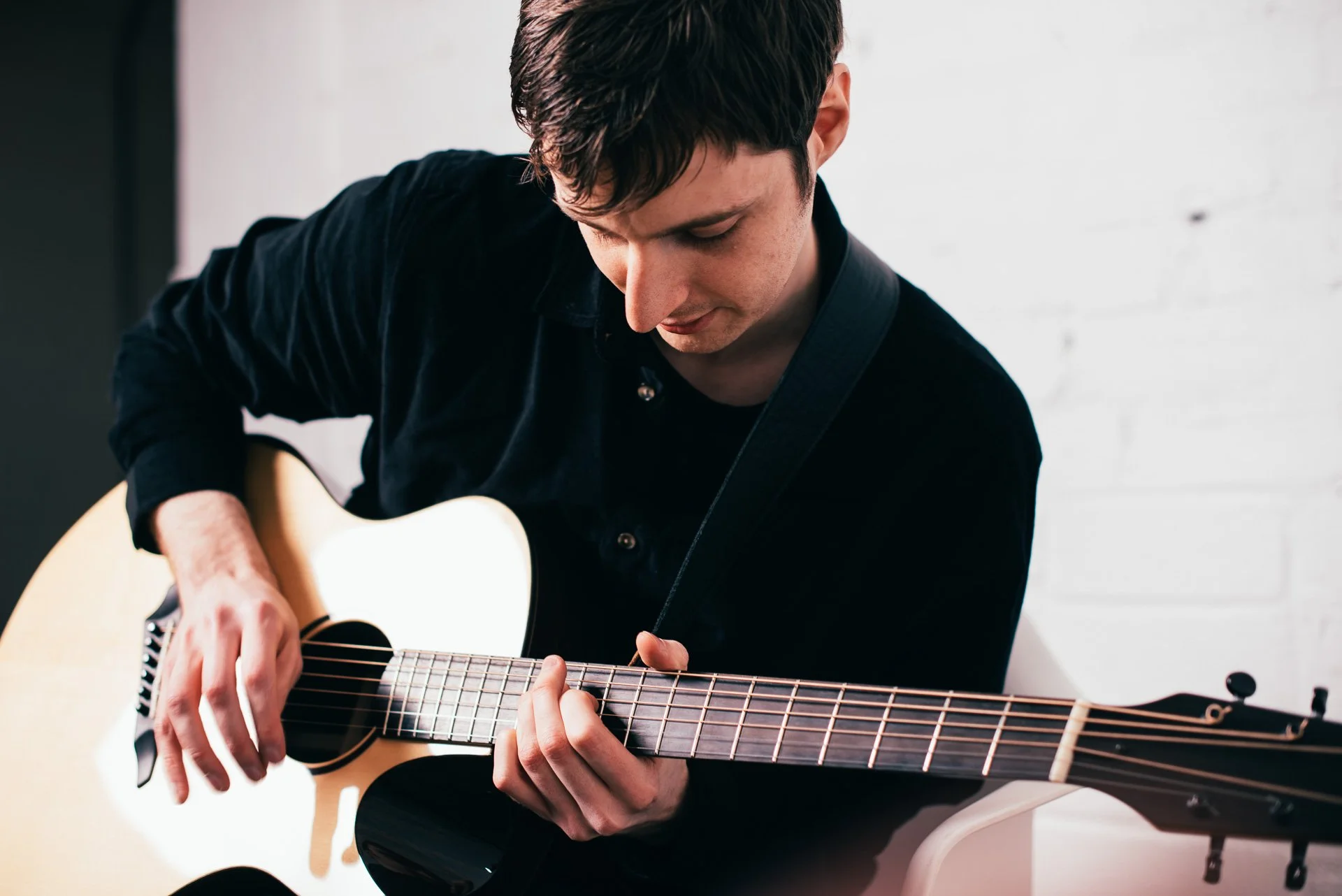 A young man playing an acoustic guitar, wearing a black shirt, sitting against a white wall.