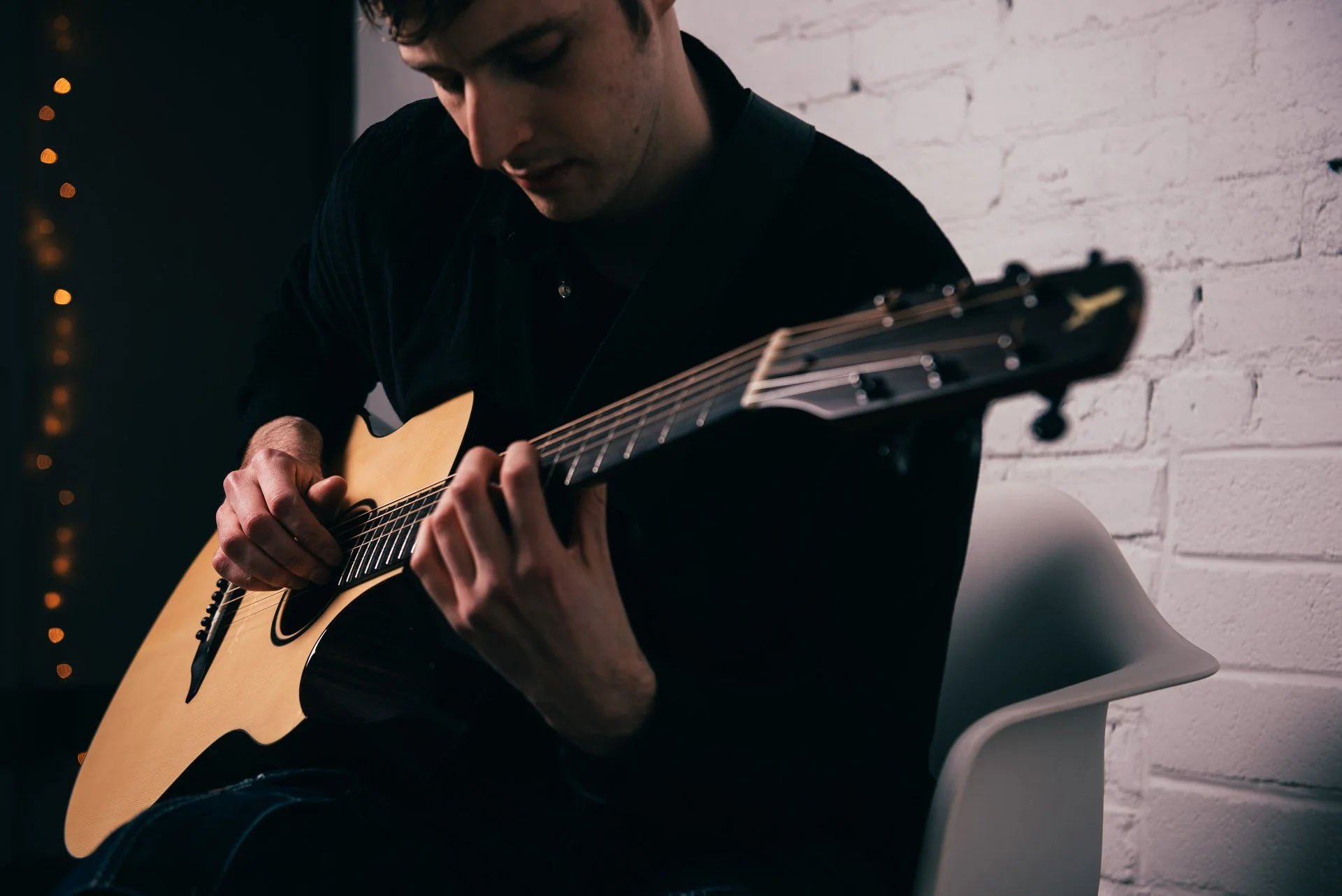 A person playing an acoustic guitar in a dimly lit room with a white brick wall in the background.