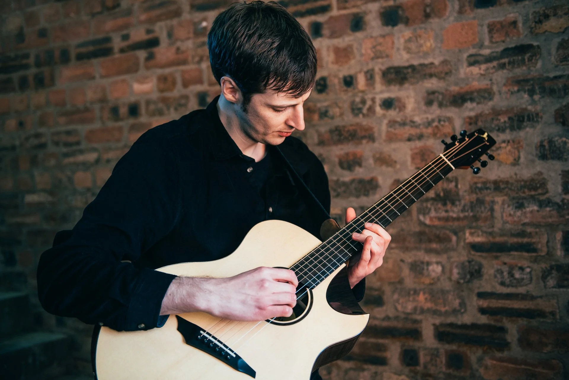 A young man playing an acoustic guitar against a brick wall.