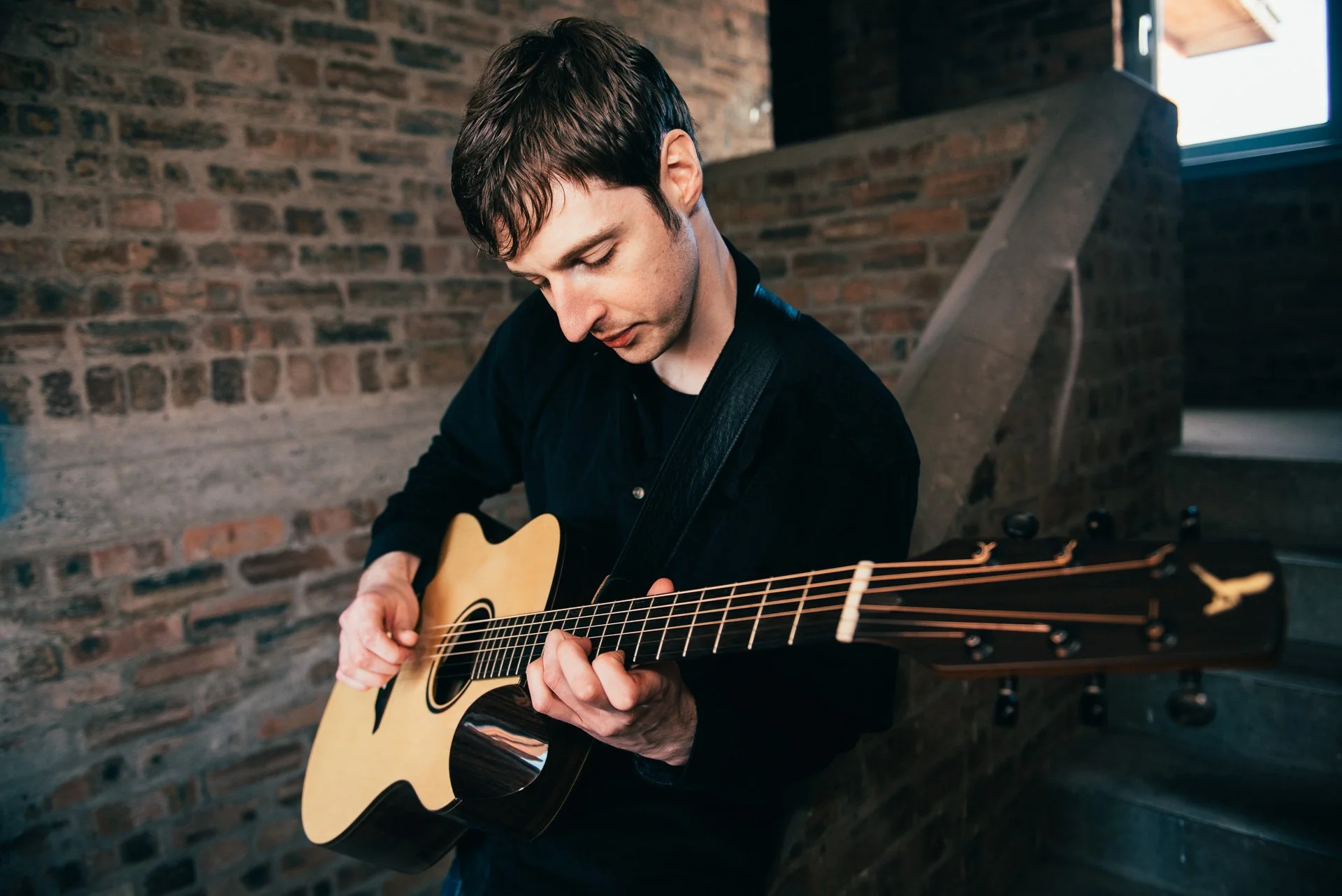 Young man with brown hair playing an acoustic guitar indoors near a brick wall and a window.