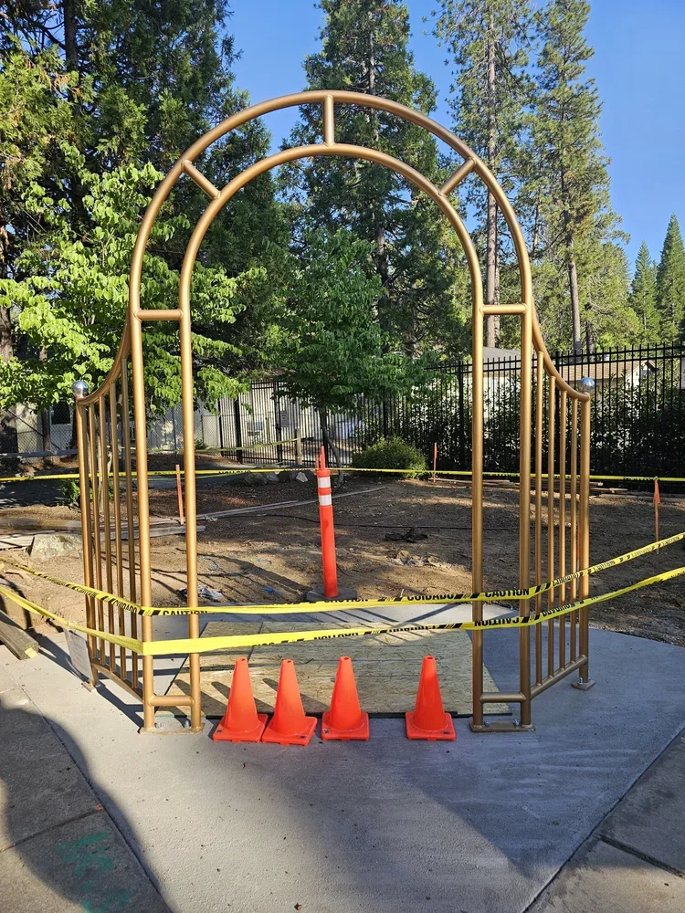 Construction site with a metal archway, orange cones, caution tape, and a traffic cone, surrounded by trees and a black fence.