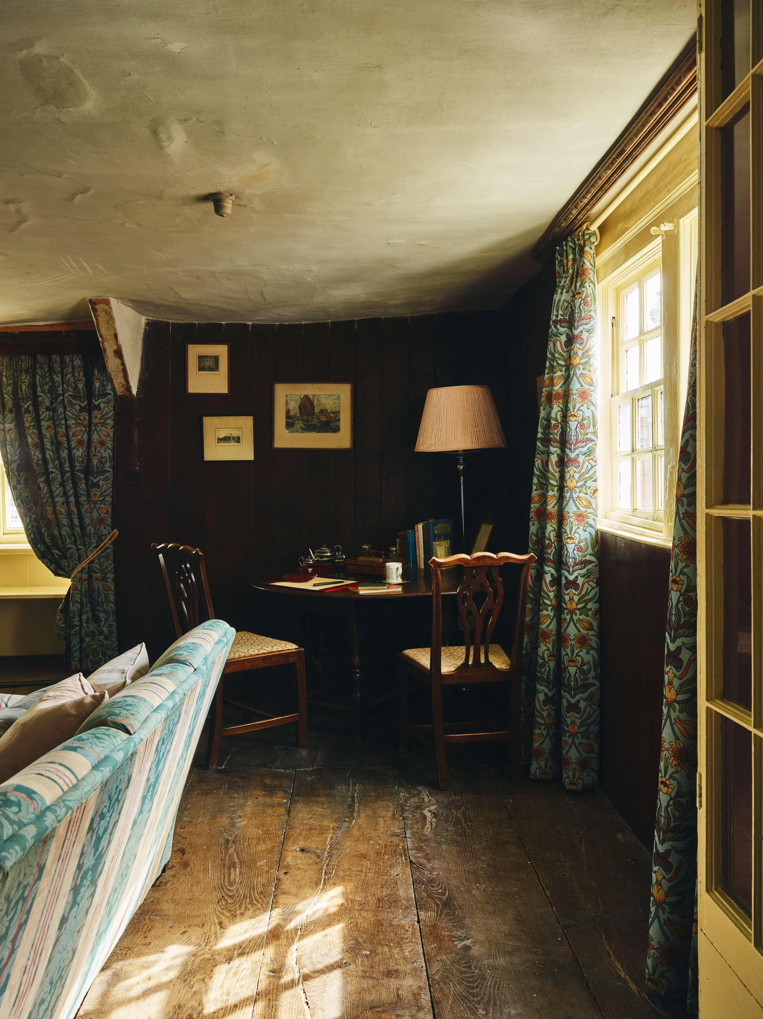 Corner of a room in a 16th century house in Cornwall with wood-paneled walls, a small desk with books and decor, two wooden chairs, a floor lamp, patterned curtains, and sunlight coming through a window.