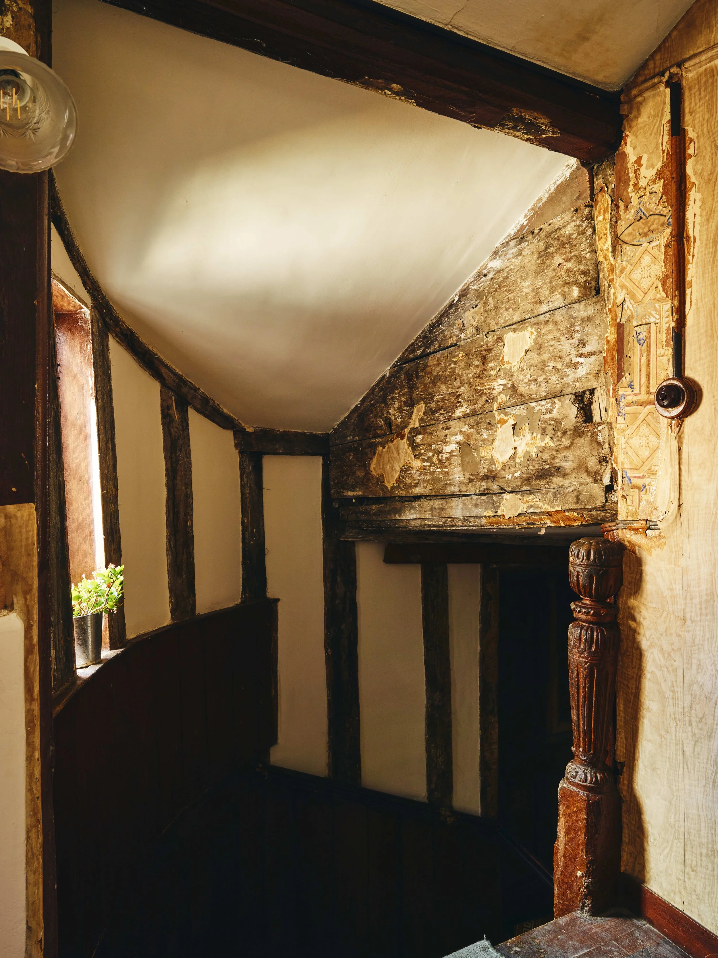 Interior of a 16th century house in Launceston with exposed wooden beams, peeling paint, and a small window with a potted plant. The staircase has a carved wooden newel post.