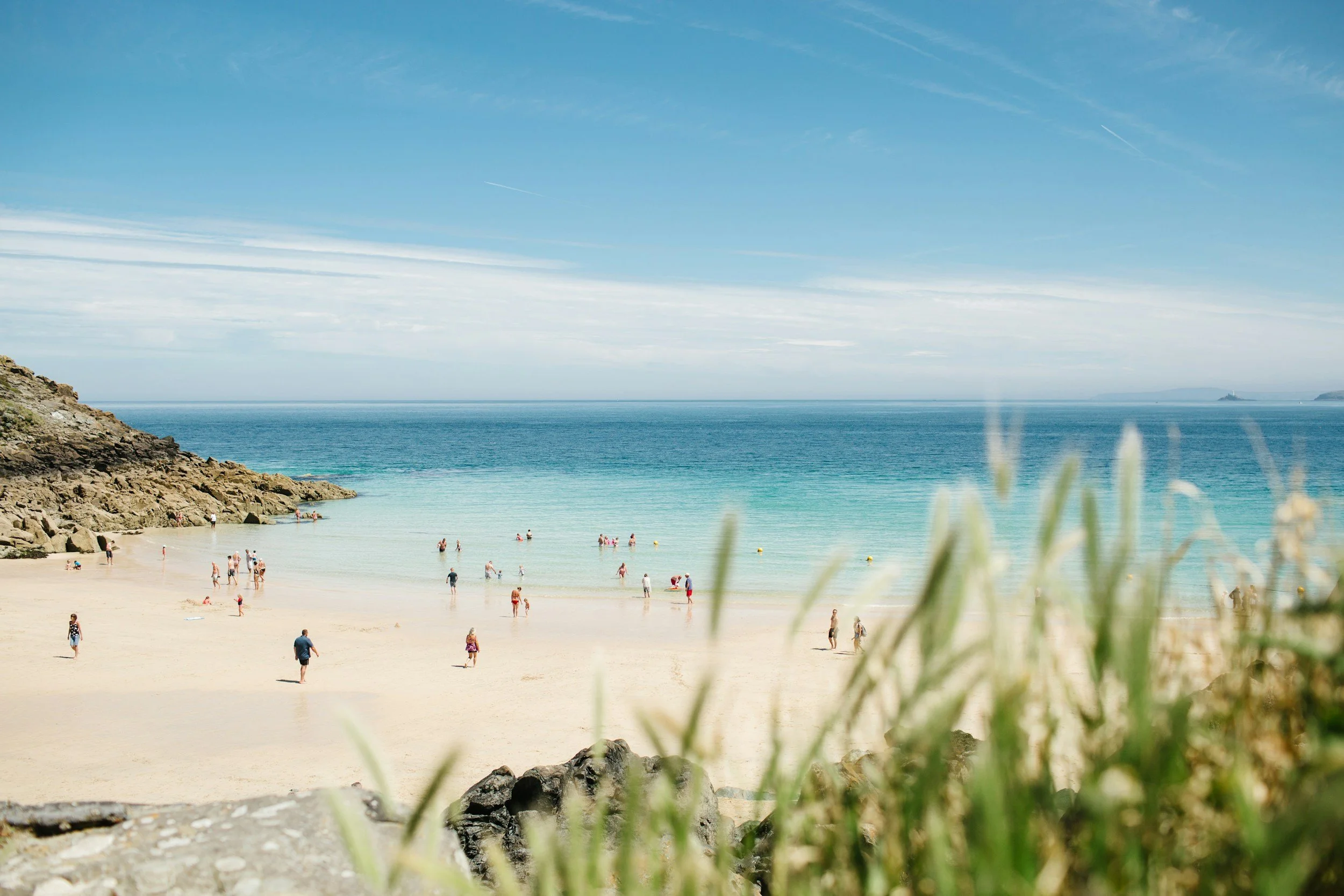 Sunny beach in Cornwall with sandy shore, people swimming and walking, rocky outcrop on the left, green foliage in the foreground, clear blue sky with thin clouds, calm ocean with a distant island on the horizon.