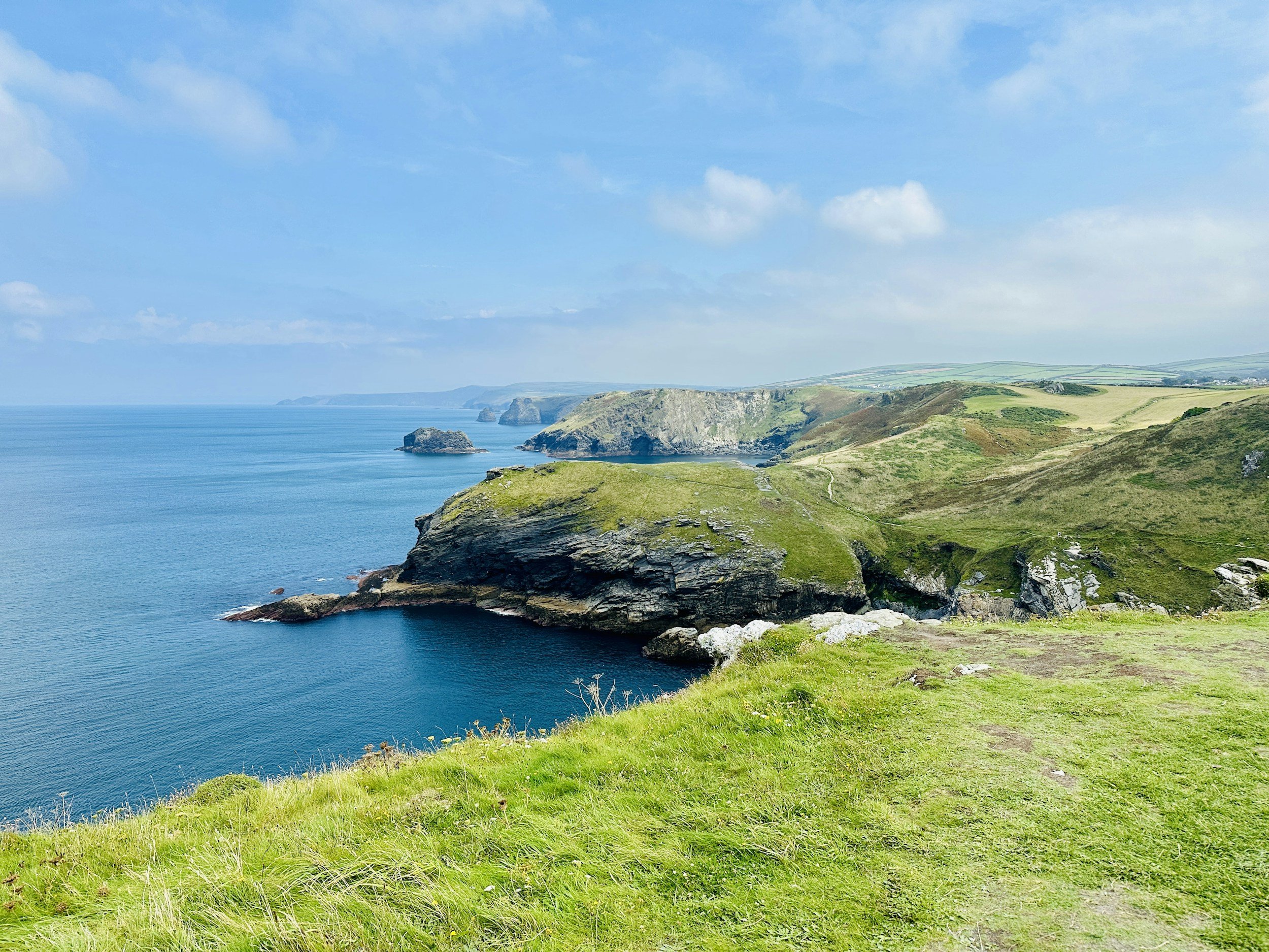 Cornish coastline with green grassy cliffs and ocean under blue sky with scattered clouds.