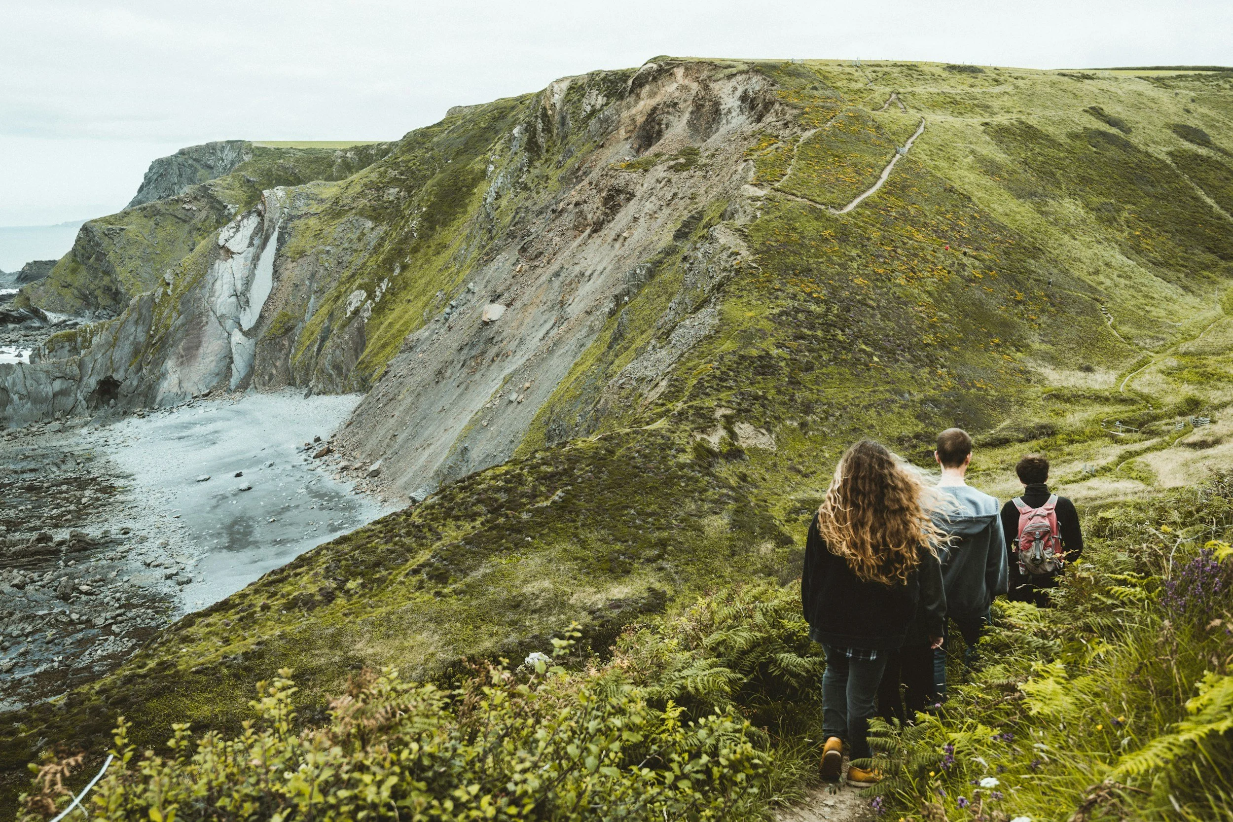 Four people hiking on a trail in Cornwall through lush green hills with cliffs and a rocky beach near the ocean.