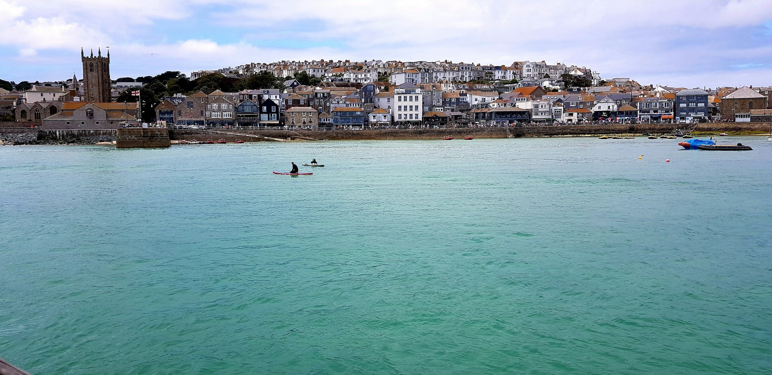 View of a Cornish town with colourful buildings along the waterfront and two people paddle boarding on calm, greenish water.