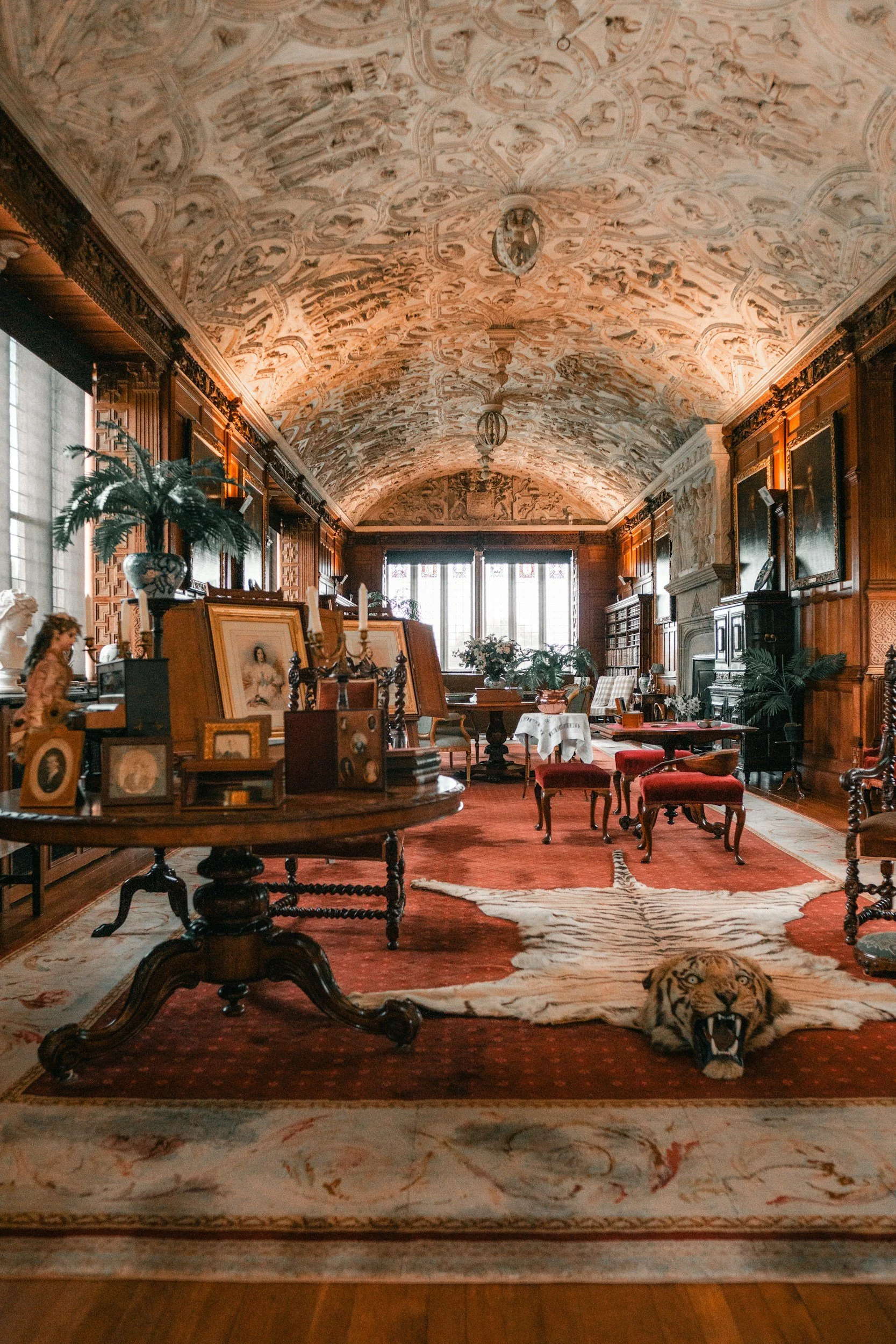 A luxurious, ornately decorated room with wooden paneling, a painted, vaulted ceiling, and a large window. The room features antique furniture, framed photographs, a tiger-striped rug with a tiger head rug lying on it, and various decorative items. There are several chairs, a table with a white cloth, potted plants, and a large fish tank visible at the far end.