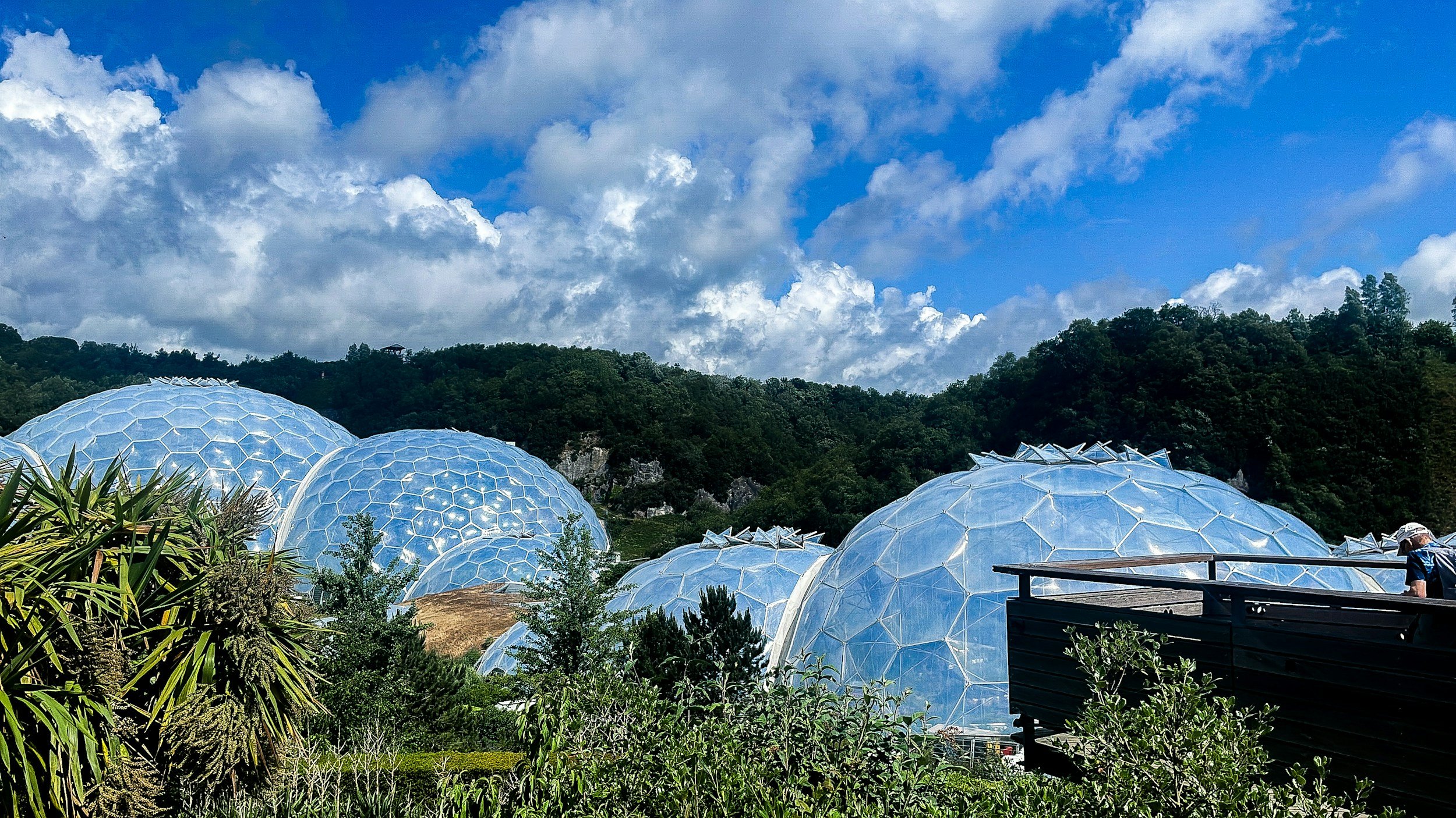 The Eden Project set in a lush green landscape with trees and hills under a partly cloudy blue sky.
