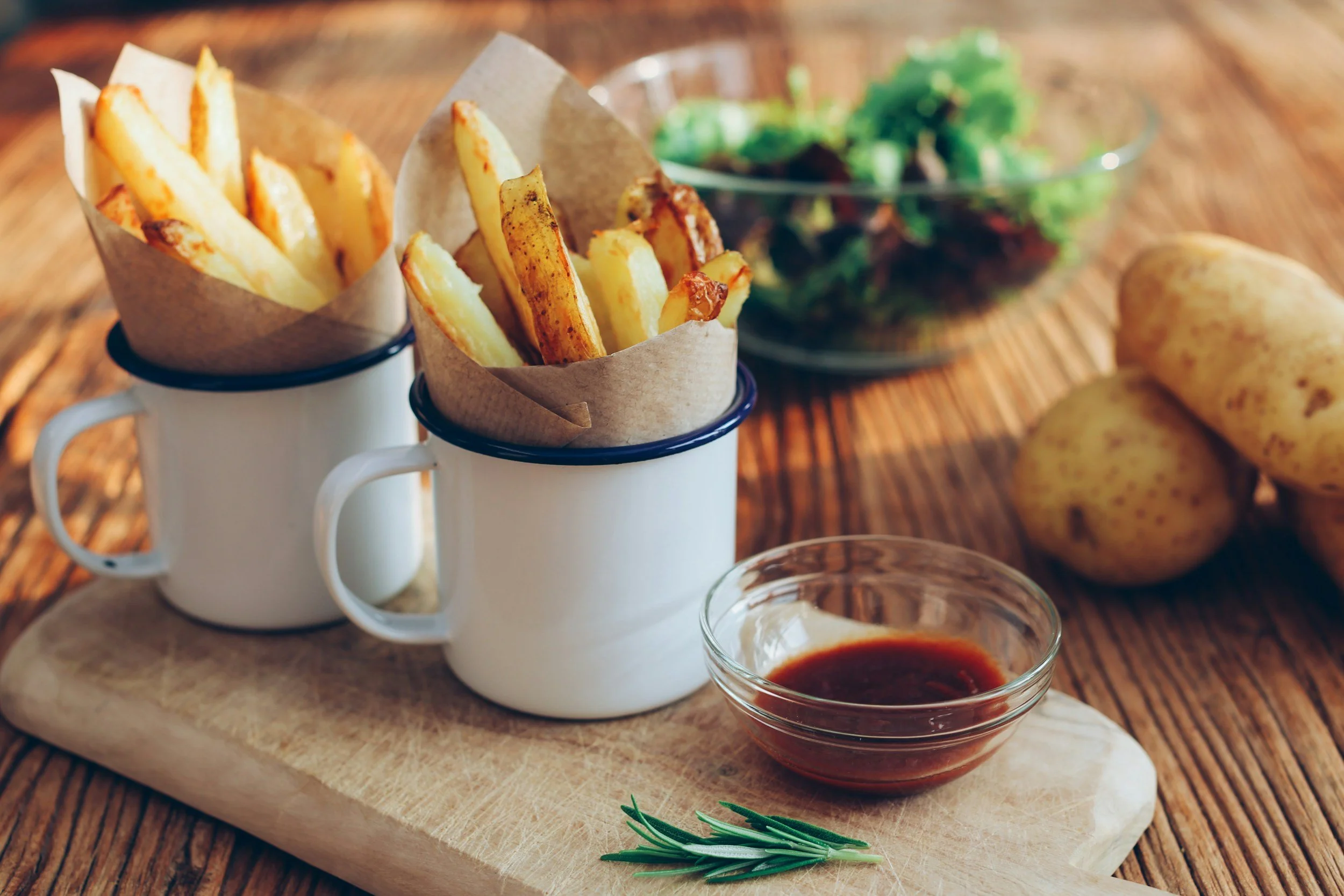 French fries in white mugs, a small bowl of dipping sauce, a sprig of rosemary, a bowl of salad, and potatoes on a wooden surface.
